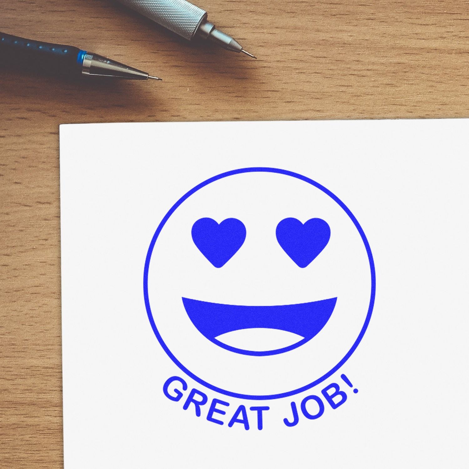 Blue heart-eyes Great Job! emoji on white paper with pens on a wooden desk, motivational success concept.