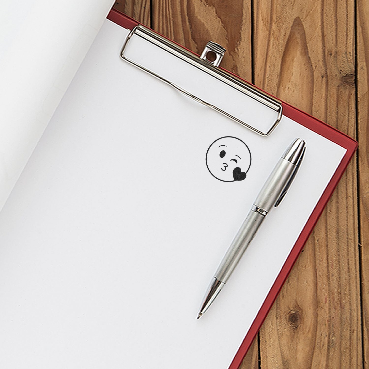 Clipboard with blank paper and silver pen on wooden desk, featuring a small kiss emoji design.