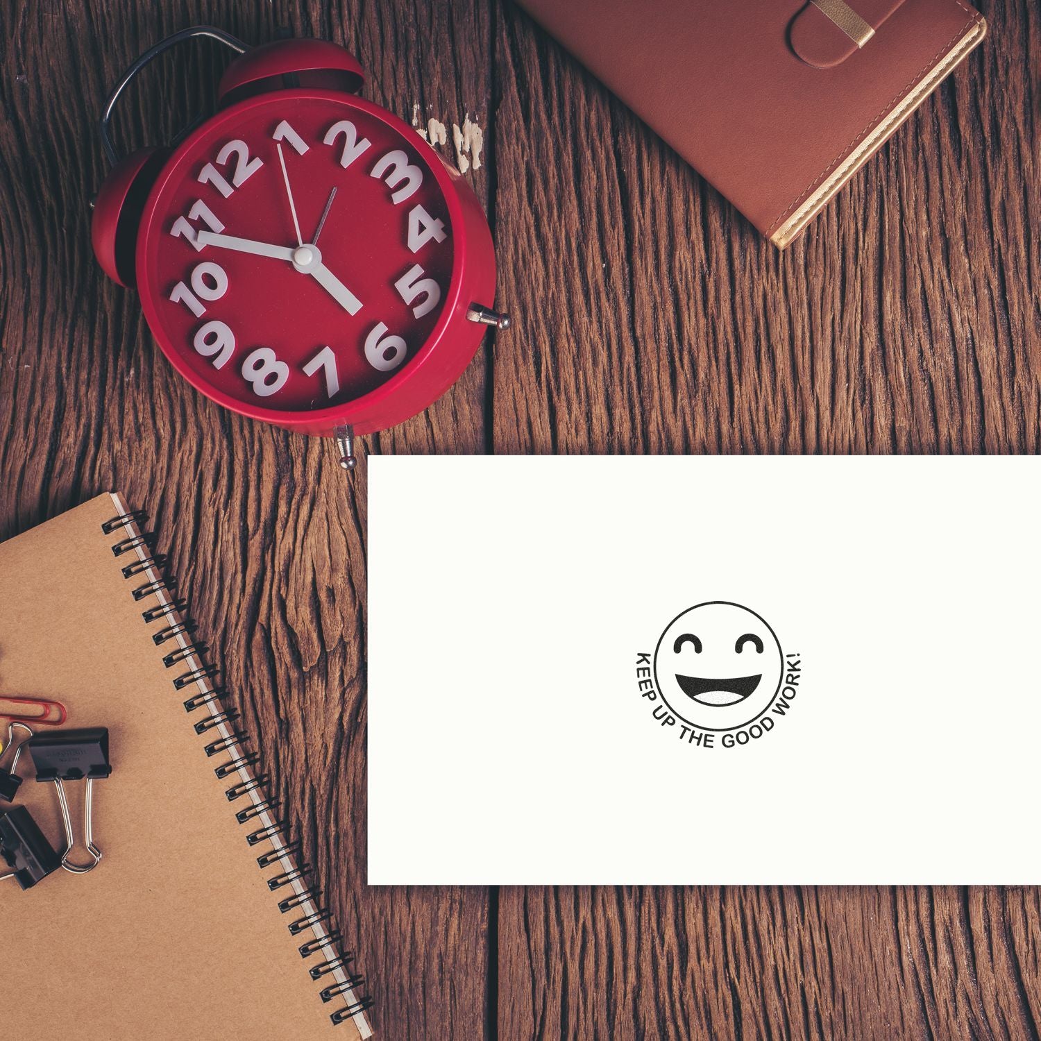 Red alarm clock, notebook, and planner on a wooden desk with a Keep Up the Good Work motivational card.