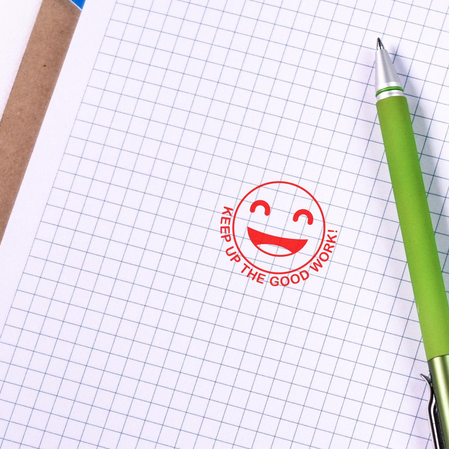 Green pen on blank graph paper notebook with Keep Up the Good Work smiley stamp, school and office stationery background.