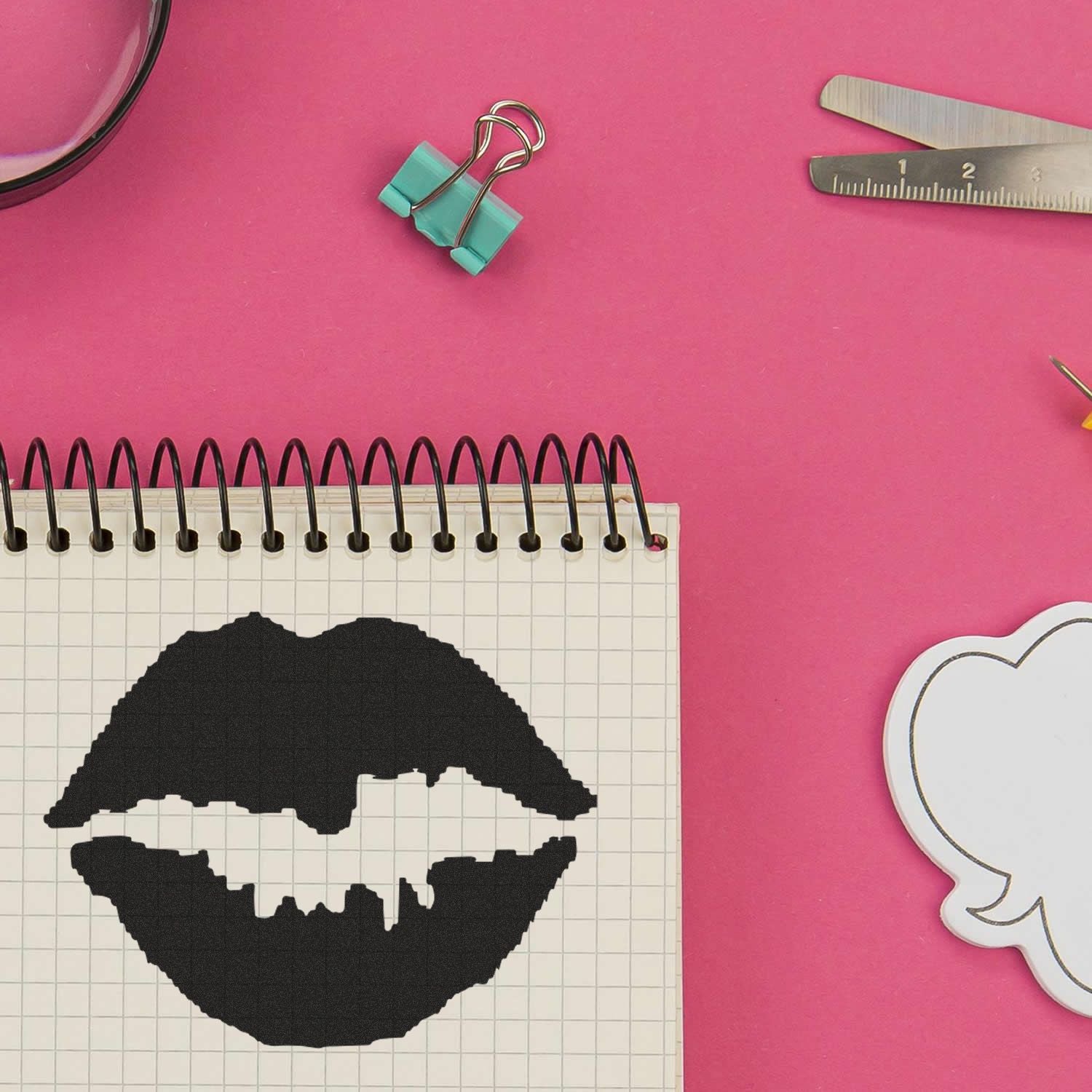 Flat lay of a notebook with black lip silhouette on a pink background, surrounded by paper clips, ruler, and craft supplies.