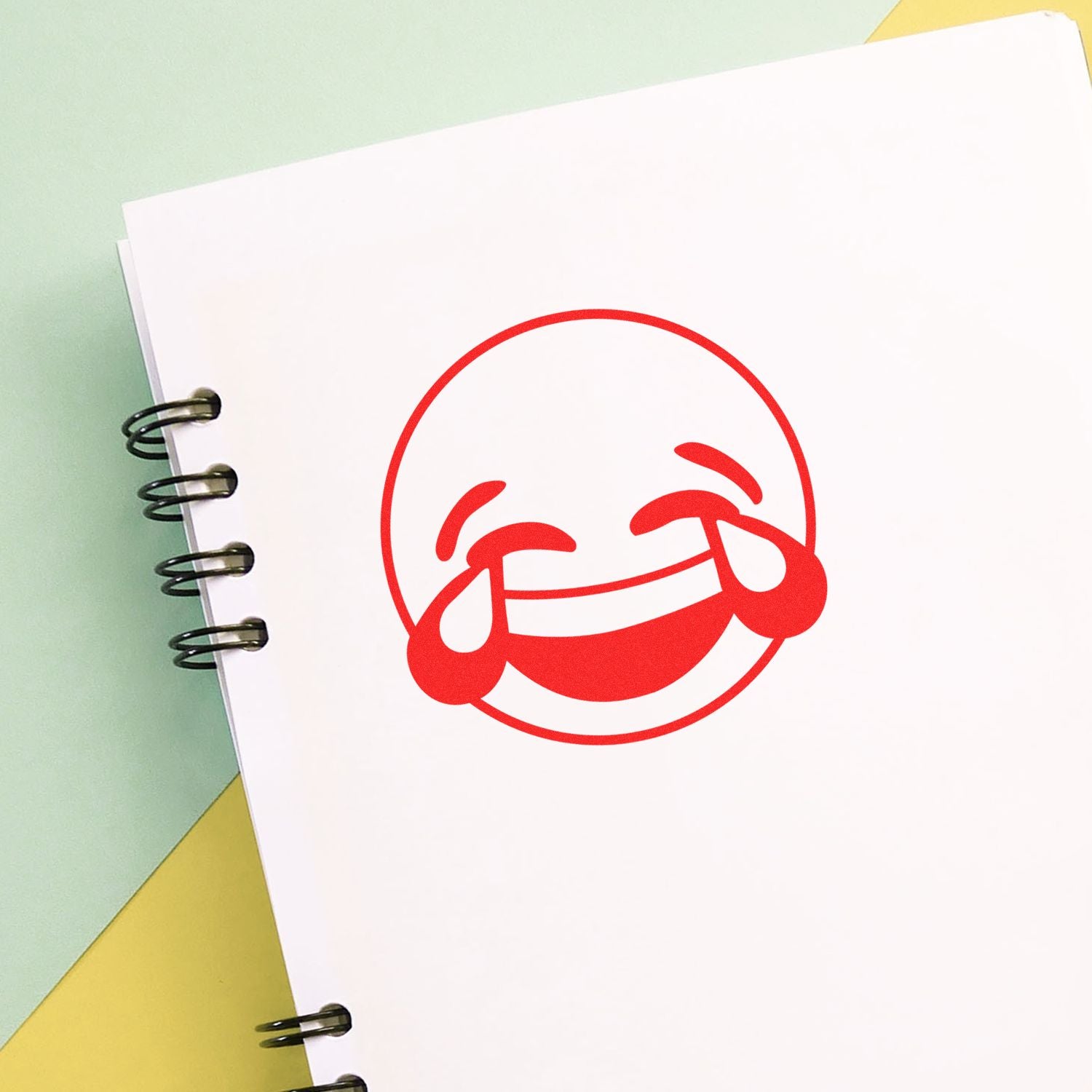 Spiral notebook with a red laughing crying emoji on a white page against a pastel background.