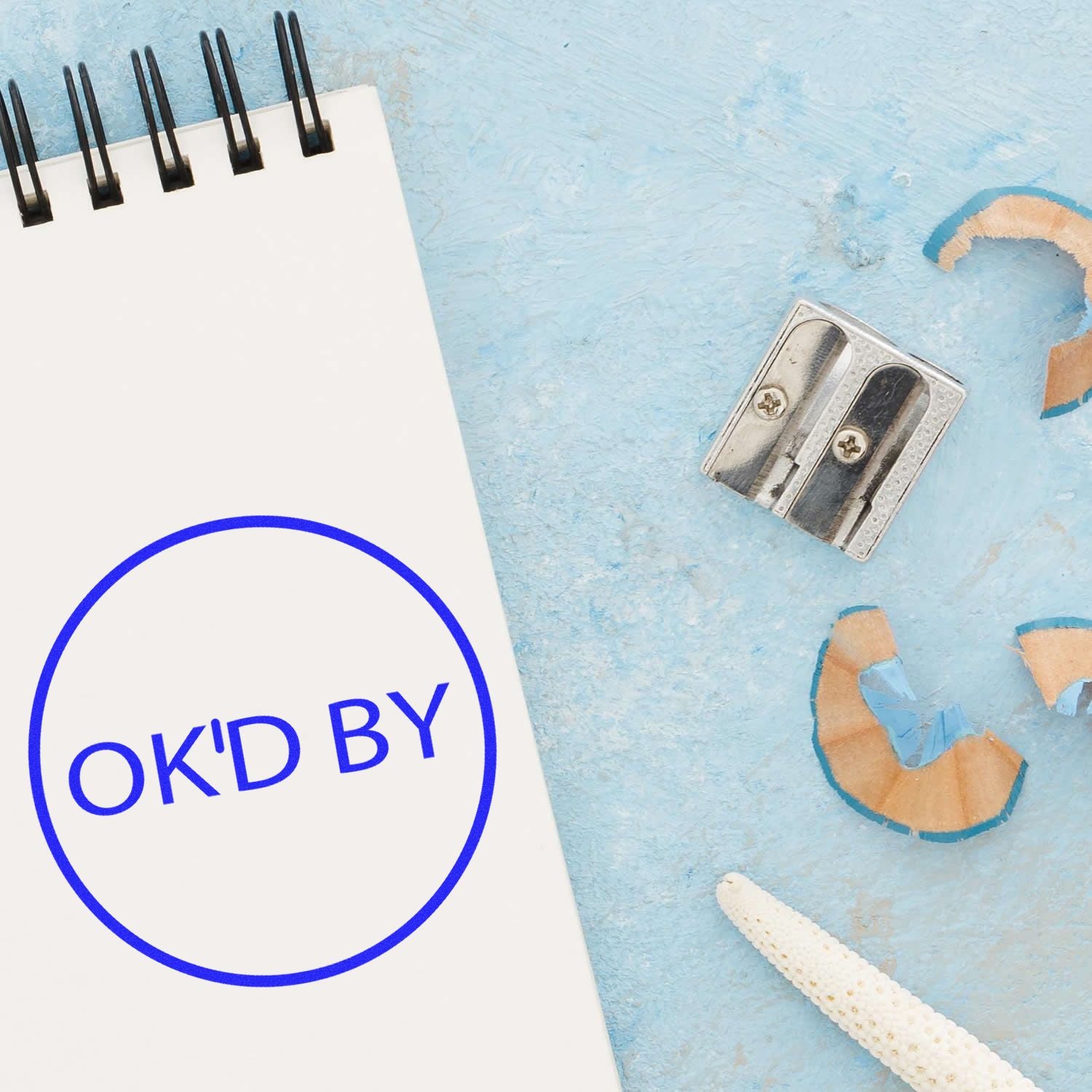 Blank notepad with OK'D BY stamp on blue background, with pencil sharpener and pencils.