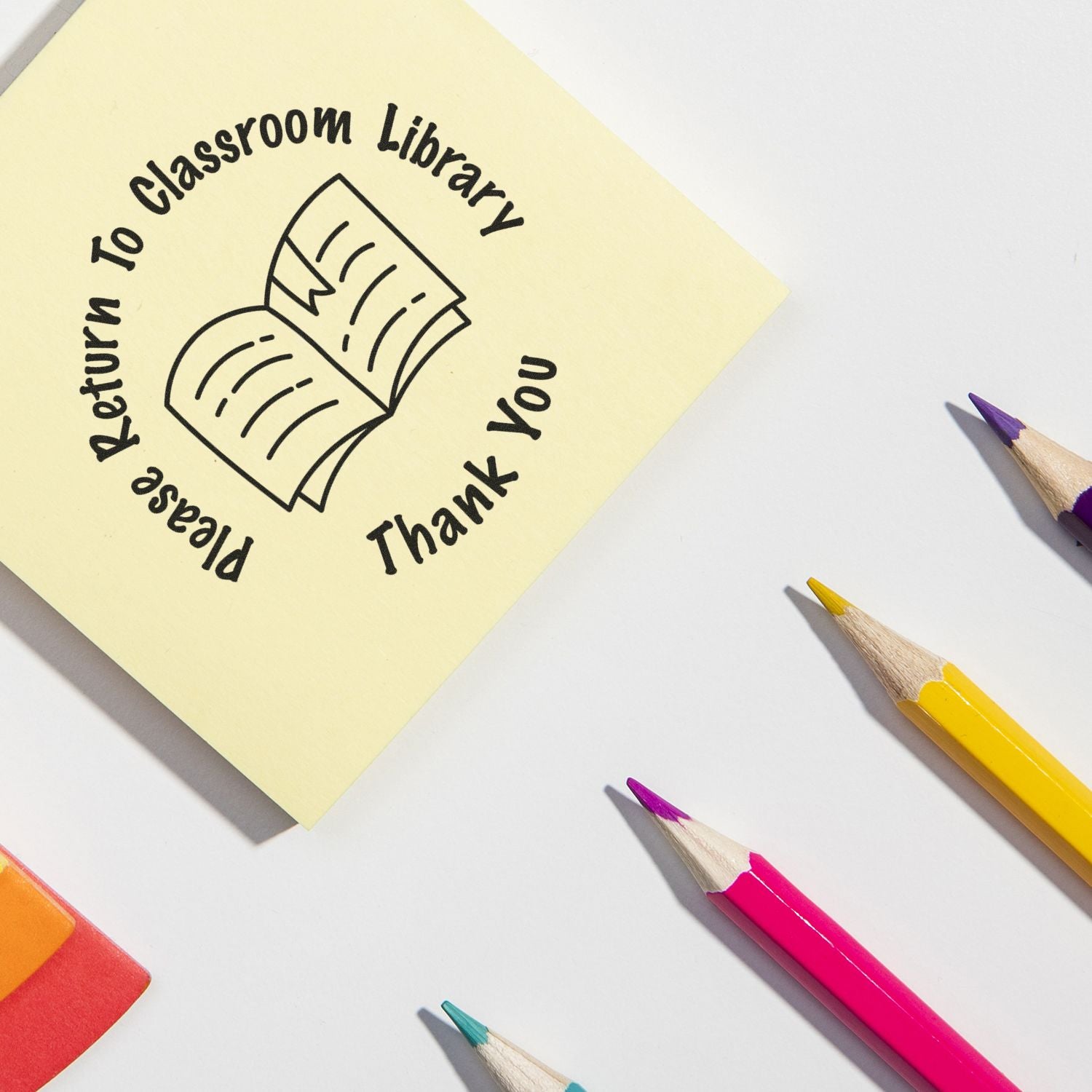 Sticky note reading Please Return to Classroom Library Thank You with open book icon and colorful pencils on a white background.