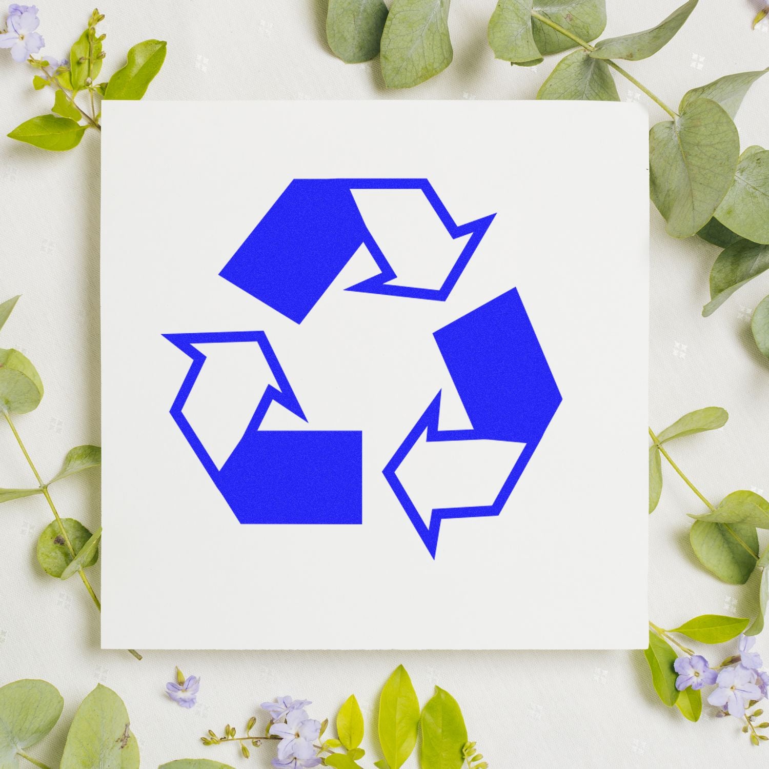 Blue recycling symbol on white paper card surrounded by green leaves and small flowers, eco-friendly sustainability concept.