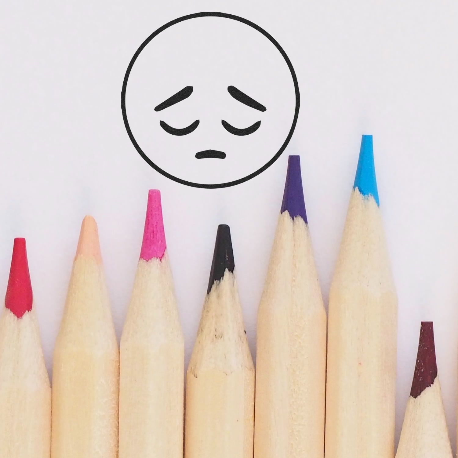 Sad face emoji above colorful colored pencil tips on a white background, symbolizing creativity and sadness.
