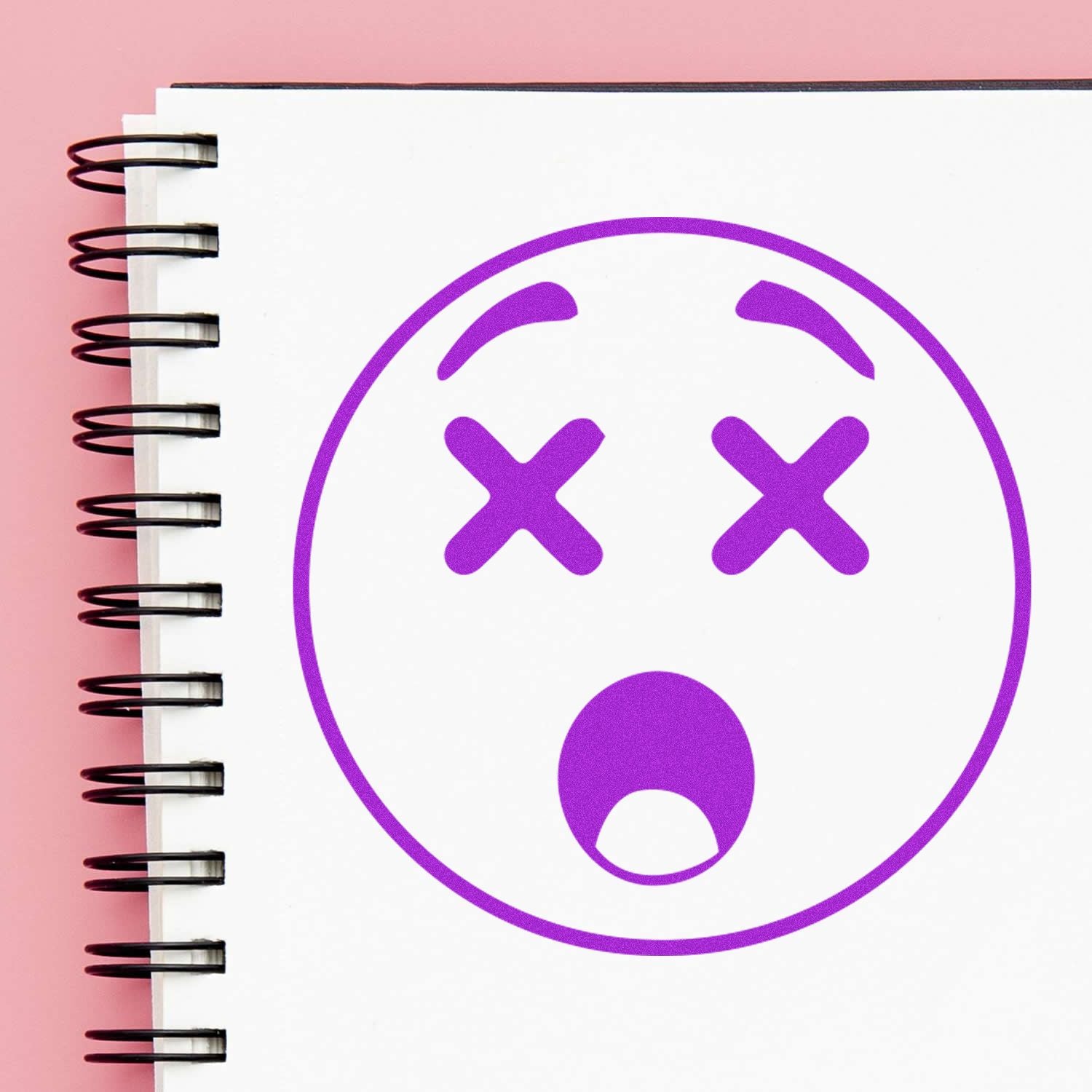 Purple dead face emoji drawn on a spiral notebook page against a pink background.