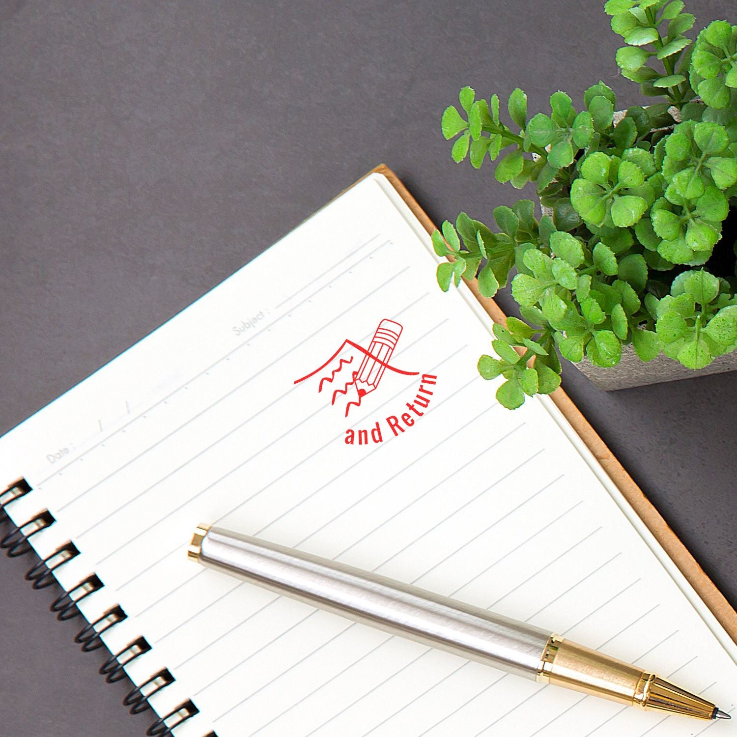 Spiral notebook with and Return stamp, silver pen, and green potted plant on dark desk background.