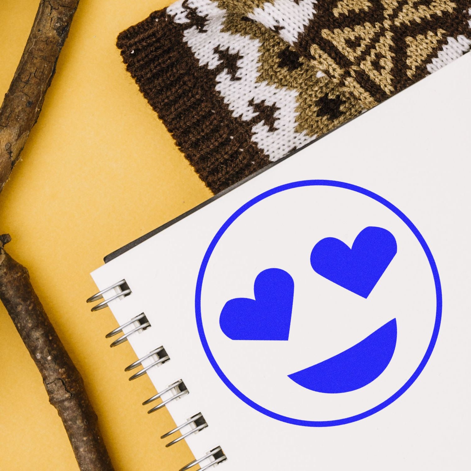 Notebook with blue heart-eyes emoji, knitted scarf, and rustic wood sticks on a yellow background.