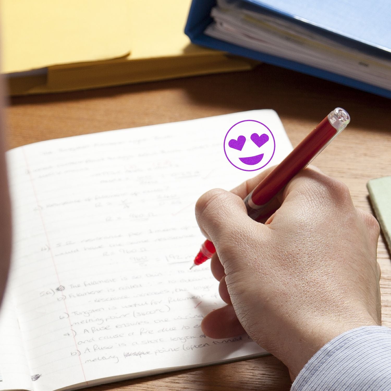 Hand writing study notes in a notebook with a red pen on a wooden desk, featuring a heart eyes emoji graphic.