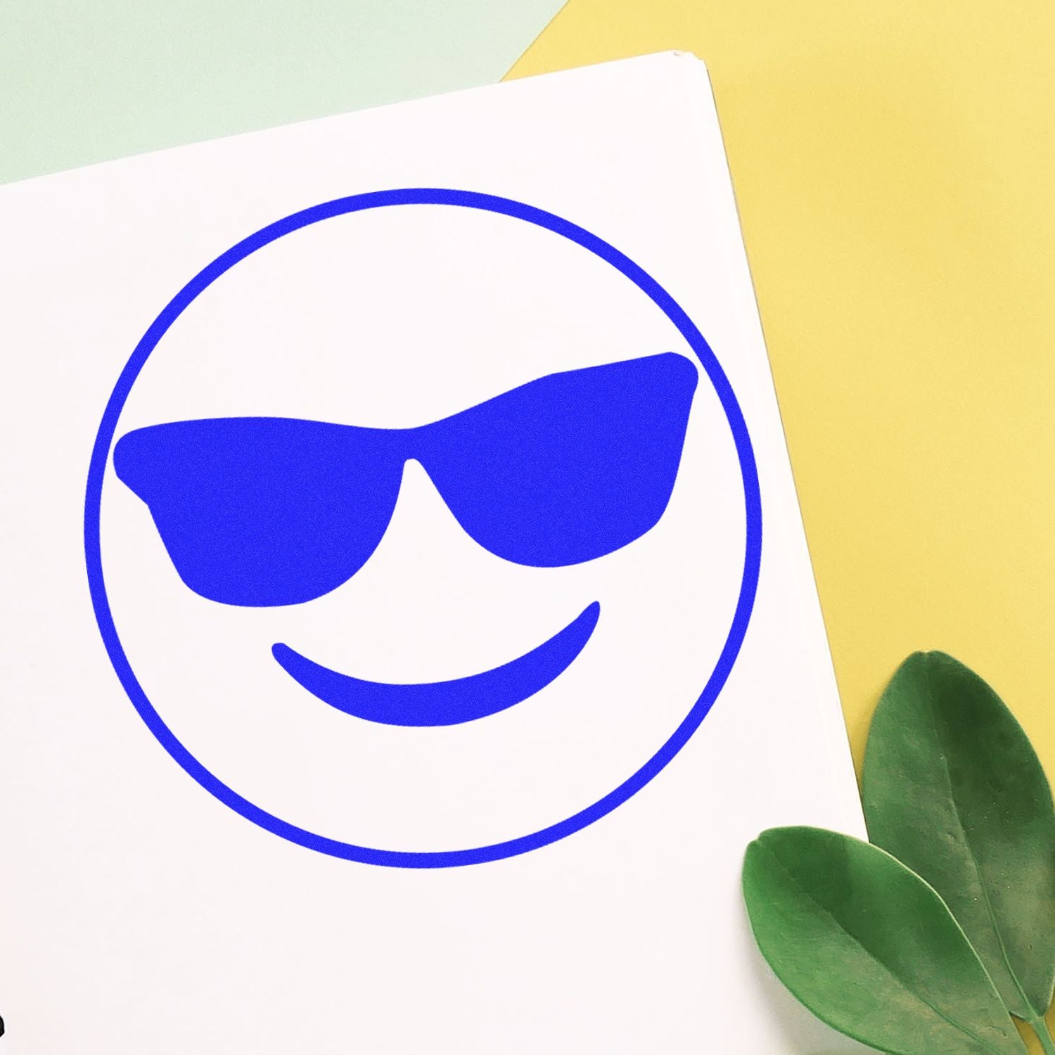 Blue sunglasses smiley face icon on white paper with yellow background and green leaves, fun modern graphic design.