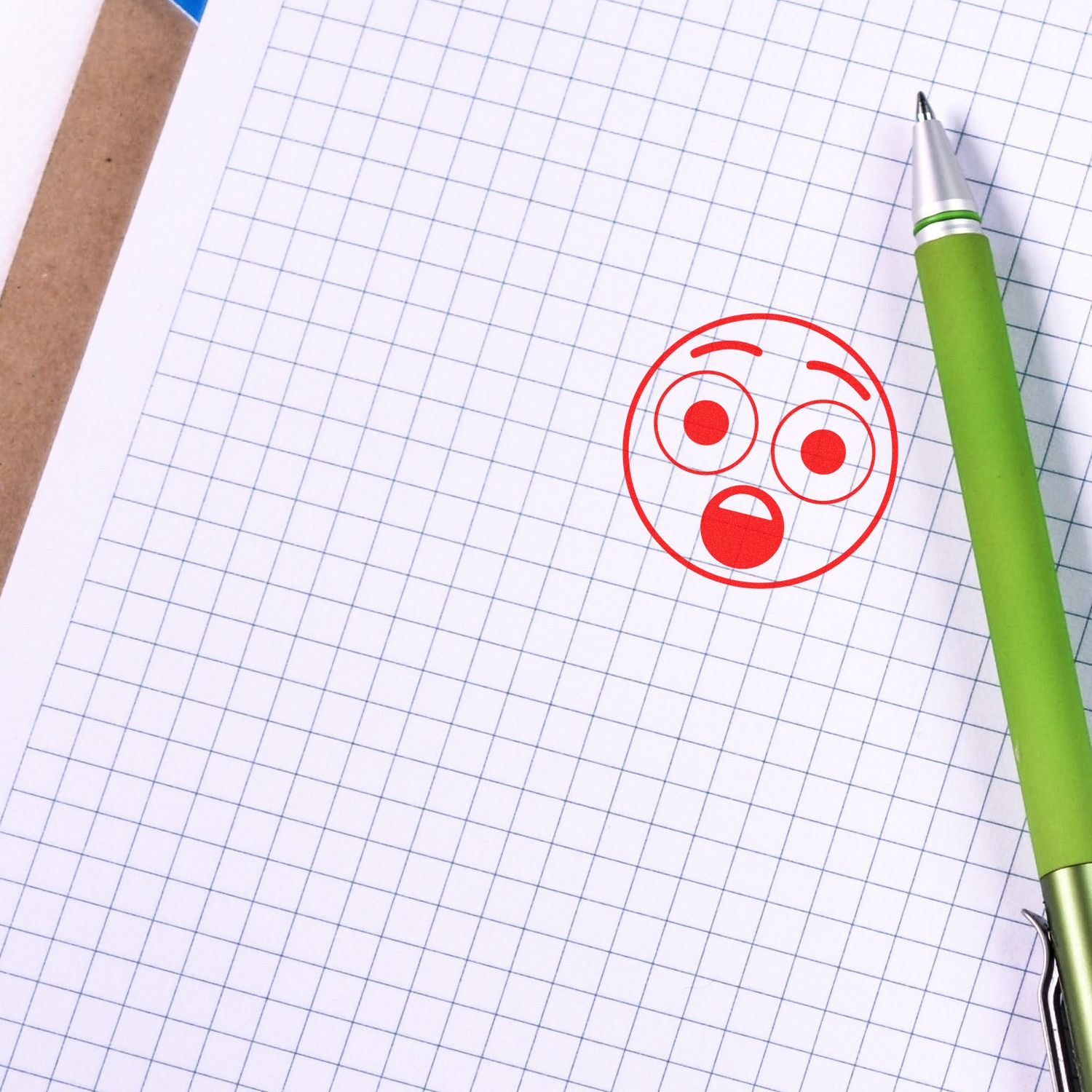 Green pen on graph paper notebook with red surprised face icon, symbolizing shock, confusion, or problem solving.