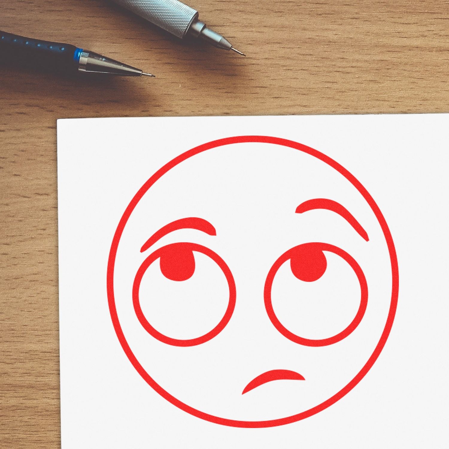 Two pens on a wooden desk beside a paper with a red sad face icon sketch