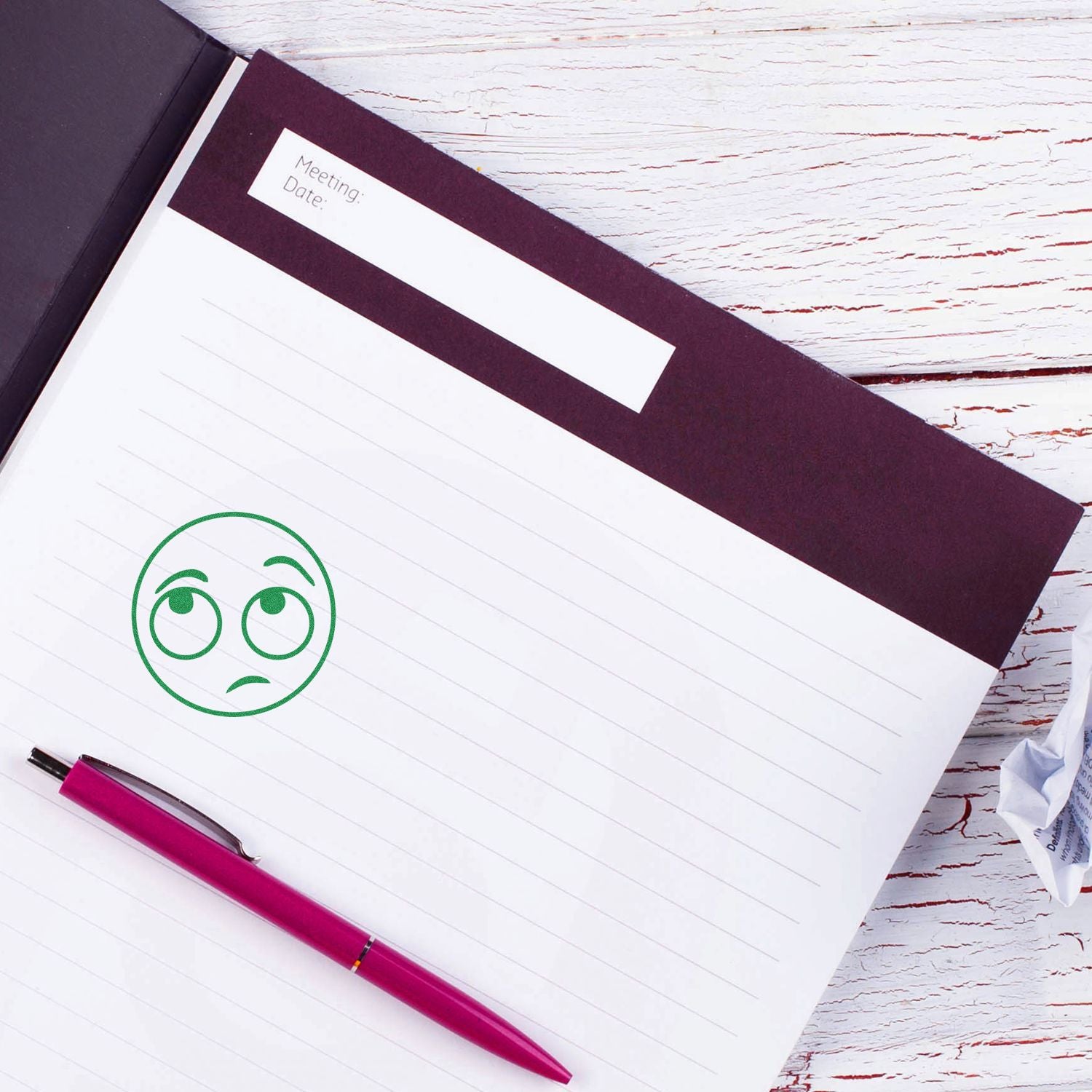 Open lined notebook with sad face doodle and pink pen on white wooden desk, meeting date page for journaling or office notes.