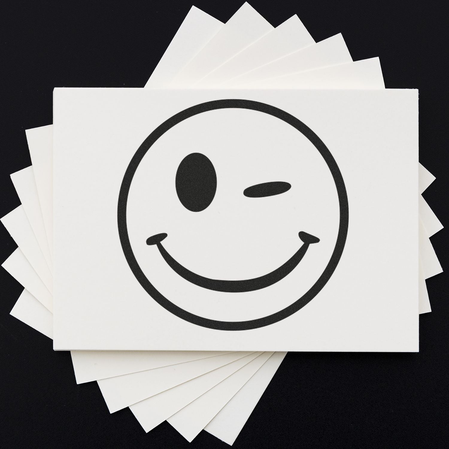 Winking smiley face icon printed on stacked white paper cards against a black background.
