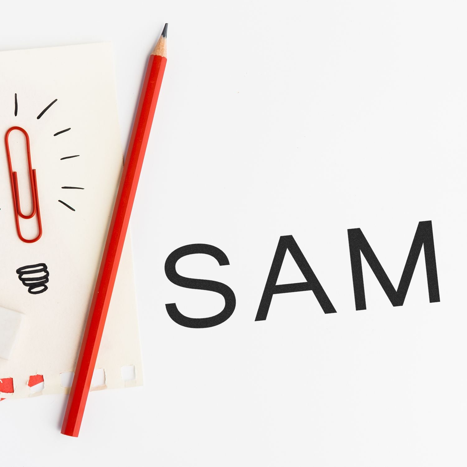 Red pencil beside a paperclip on a white notebook with the word SAM in bold black letters.