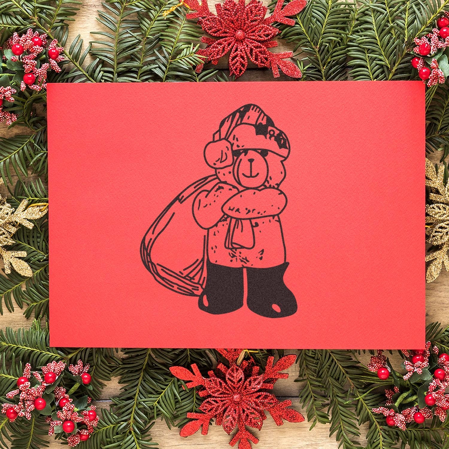 Festive Christmas teddy bear card surrounded by evergreen garland and red holiday decorations.