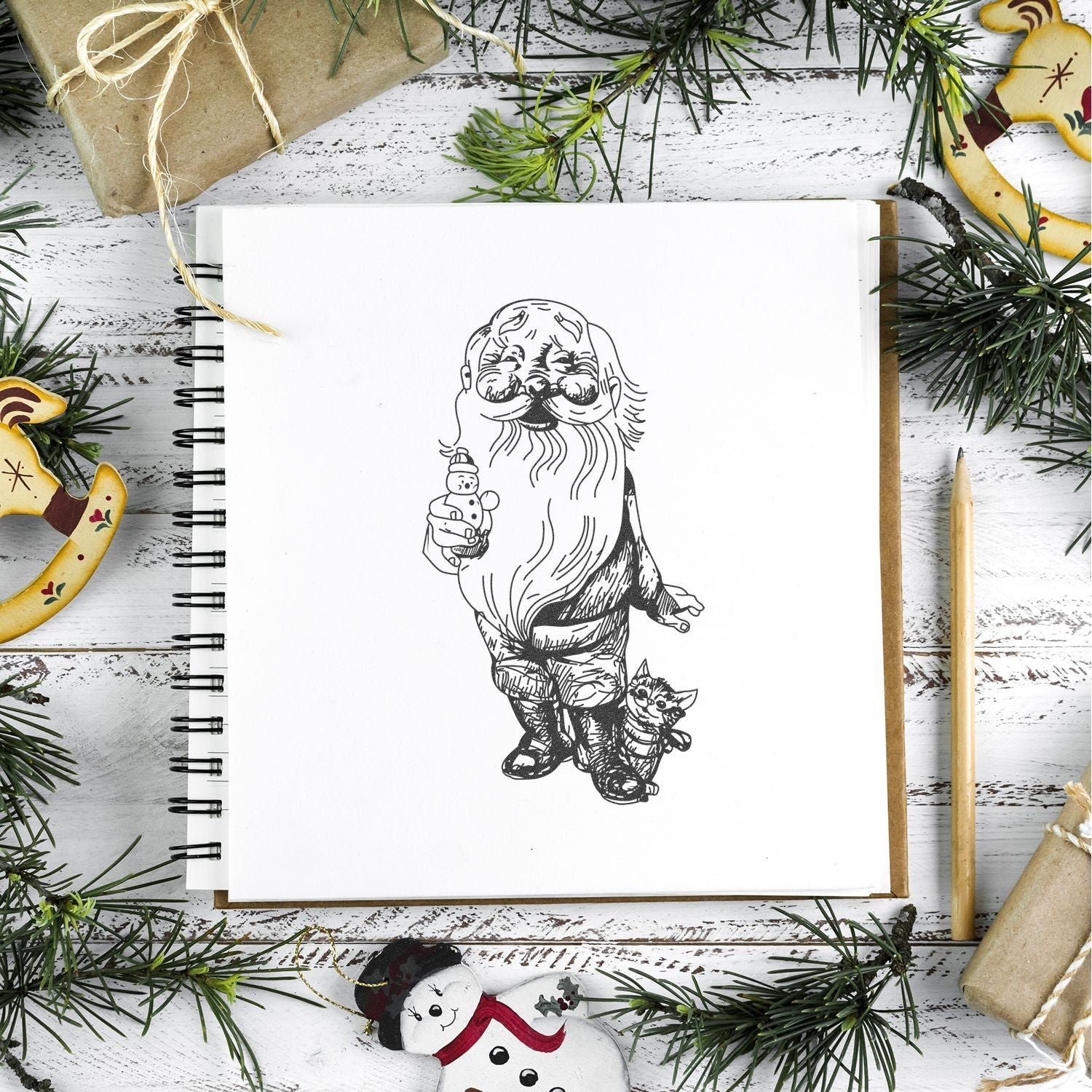 Sketchbook with vintage Santa Claus drawing surrounded by Christmas decorations, pine branches, and wrapped gifts on rustic white wood background