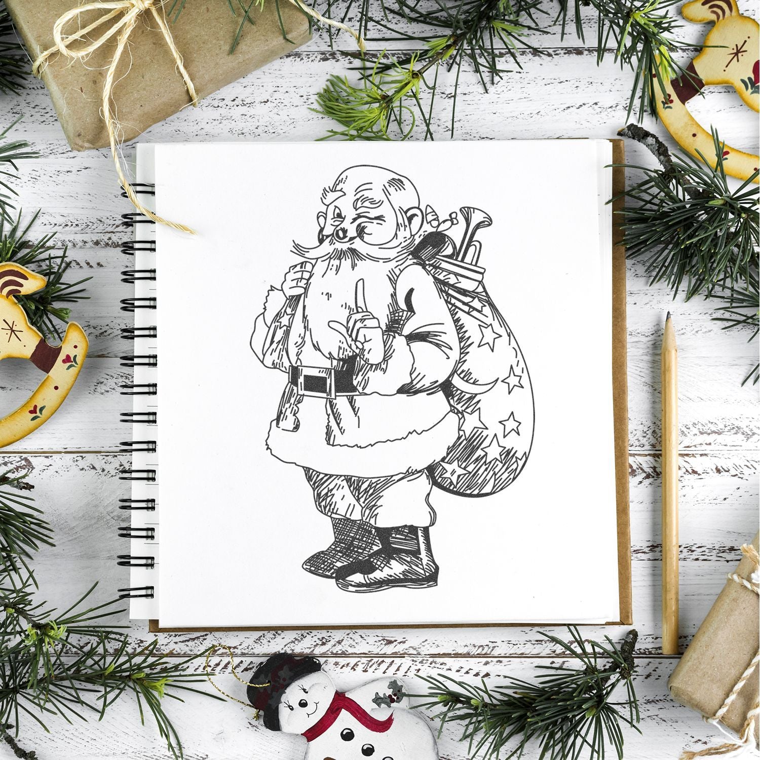 Hand-drawn Santa Claus illustration in a sketchbook surrounded by Christmas decorations and pine branches on a rustic white wooden background.