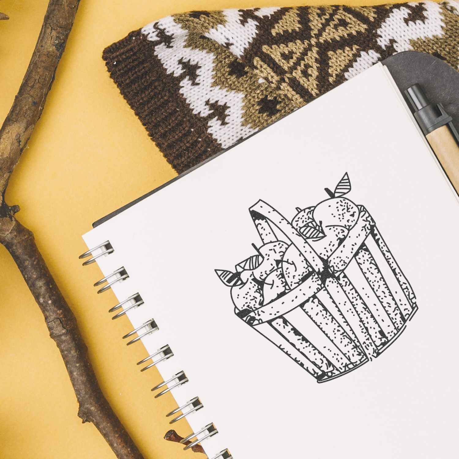 Sketchbook with hand-drawn cupcake illustration, knitted pouch, pen, and rustic twigs on a yellow background.