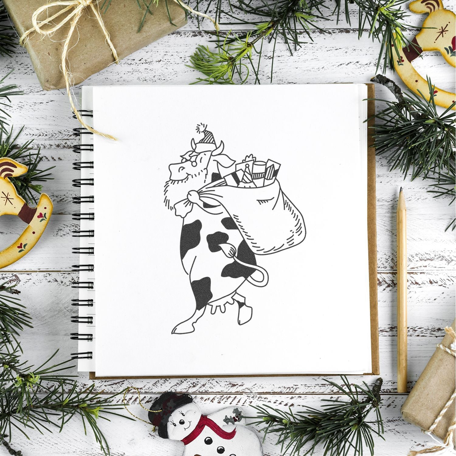 Festive holiday flat lay with a sketchbook showing a cow carrying Christmas gifts, surrounded by pine branches, wrapped presents, and rustic decorations.