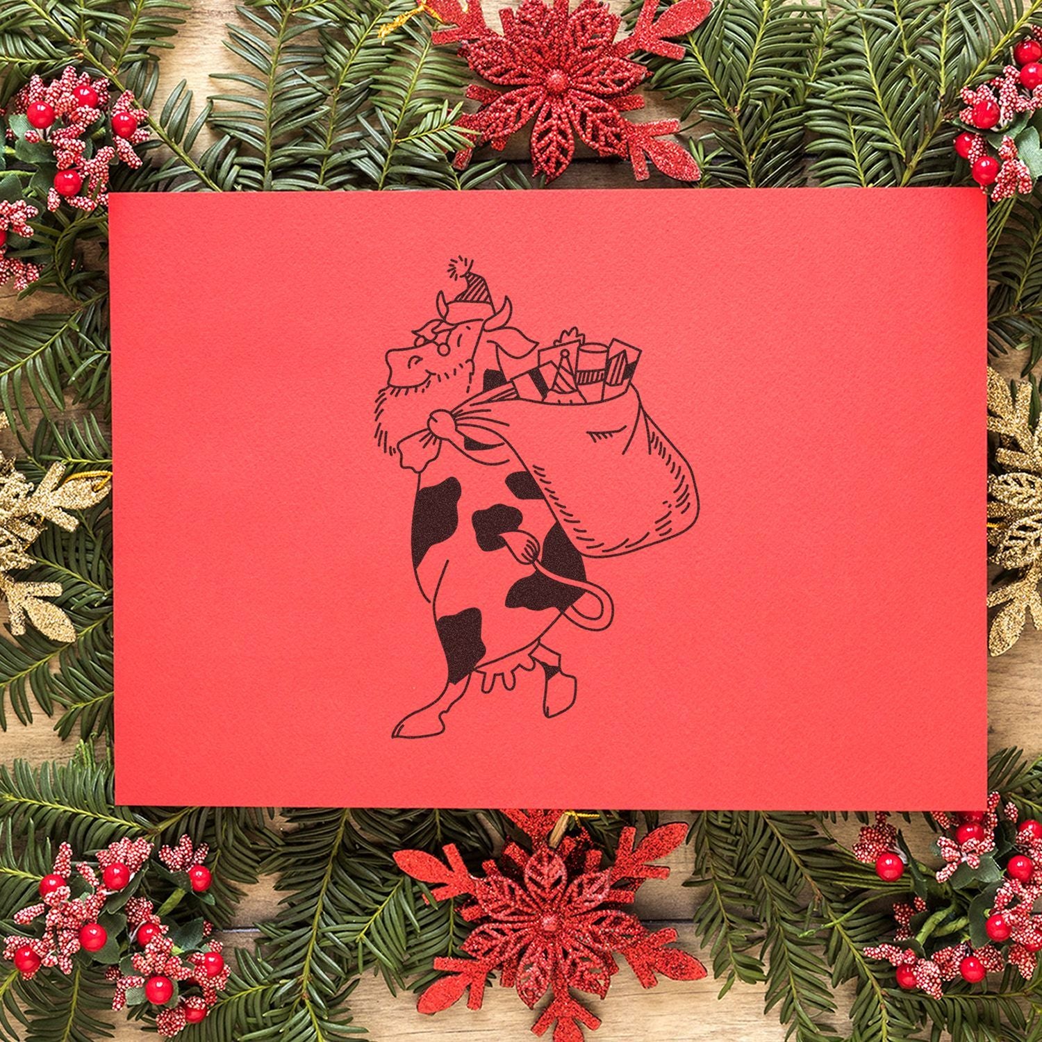 Festive red holiday card with cartoon cow carrying gifts, framed by evergreen branches and red Christmas decorations on a rustic wooden background.