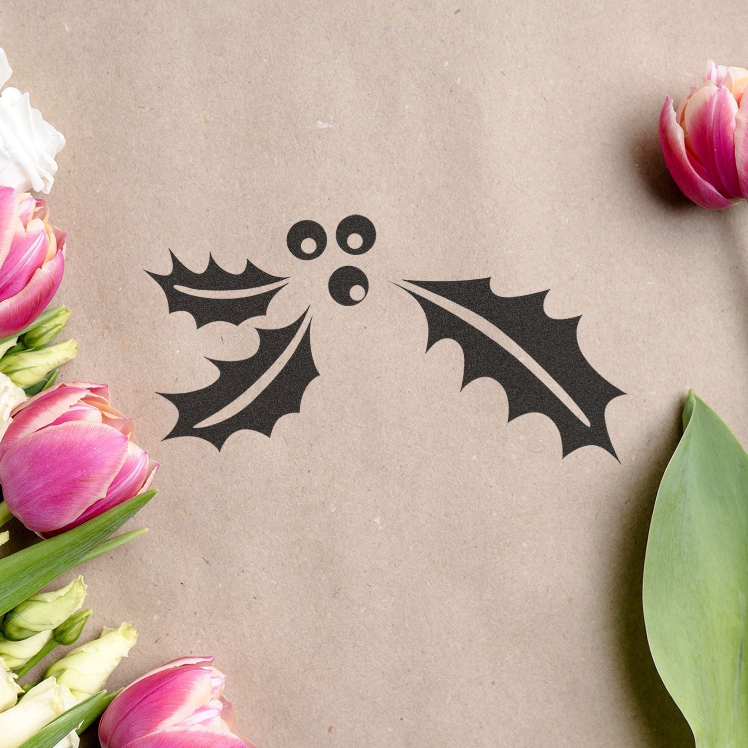 Black holly leaf and berry holiday design on kraft paper, framed by pink tulips and green leaves, floral seasonal background.
