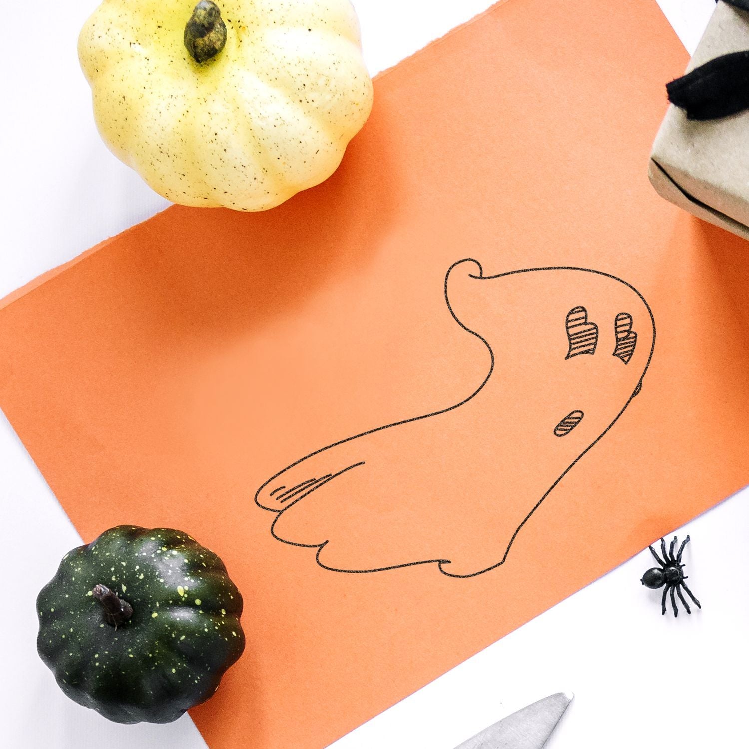 Halloween craft flat lay with orange paper ghost outline, decorative pumpkins, and a plastic spider on a white background