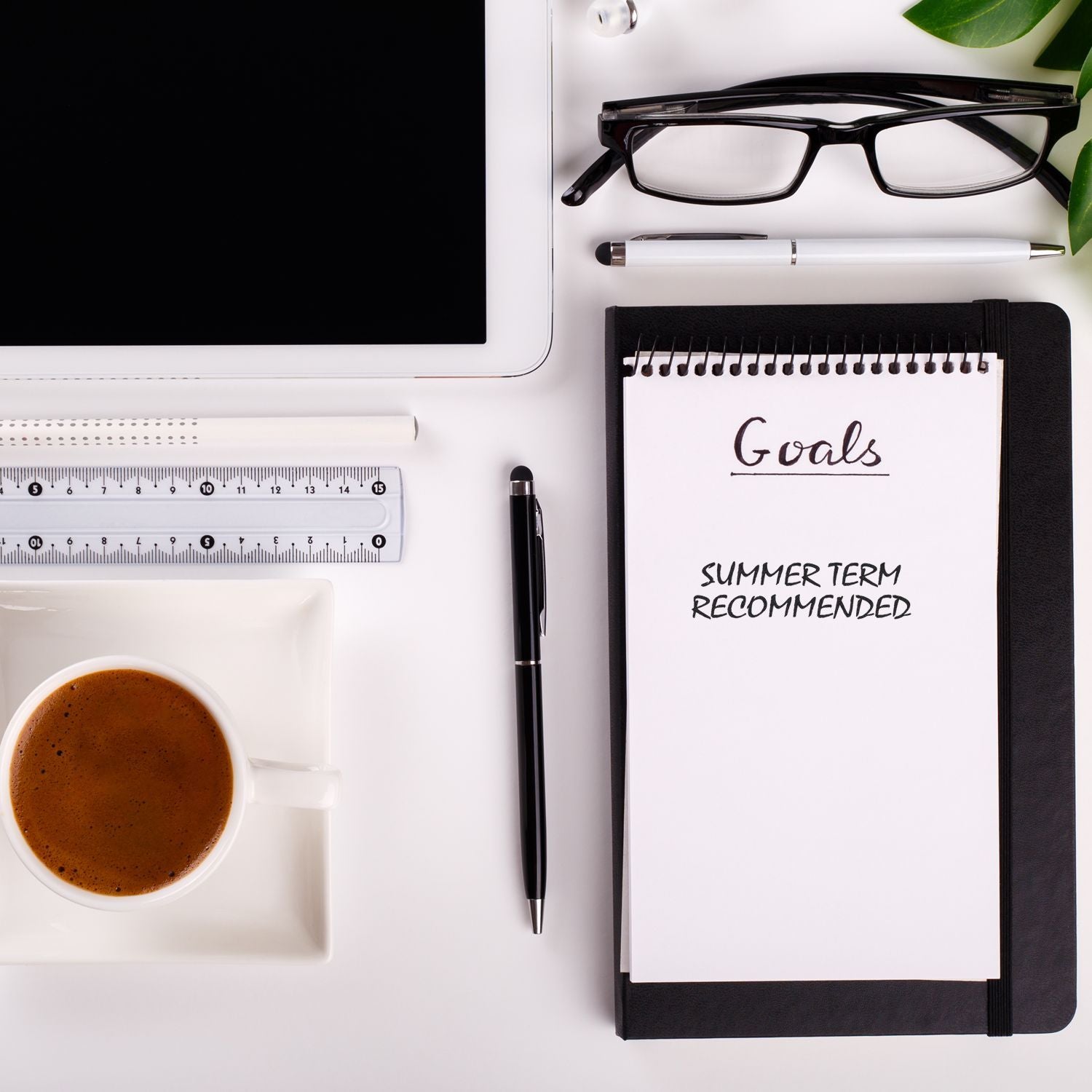 Desk flat lay with Goals notebook reading Summer Term Recommended, glasses, pen, and coffee for summer study goal planning.