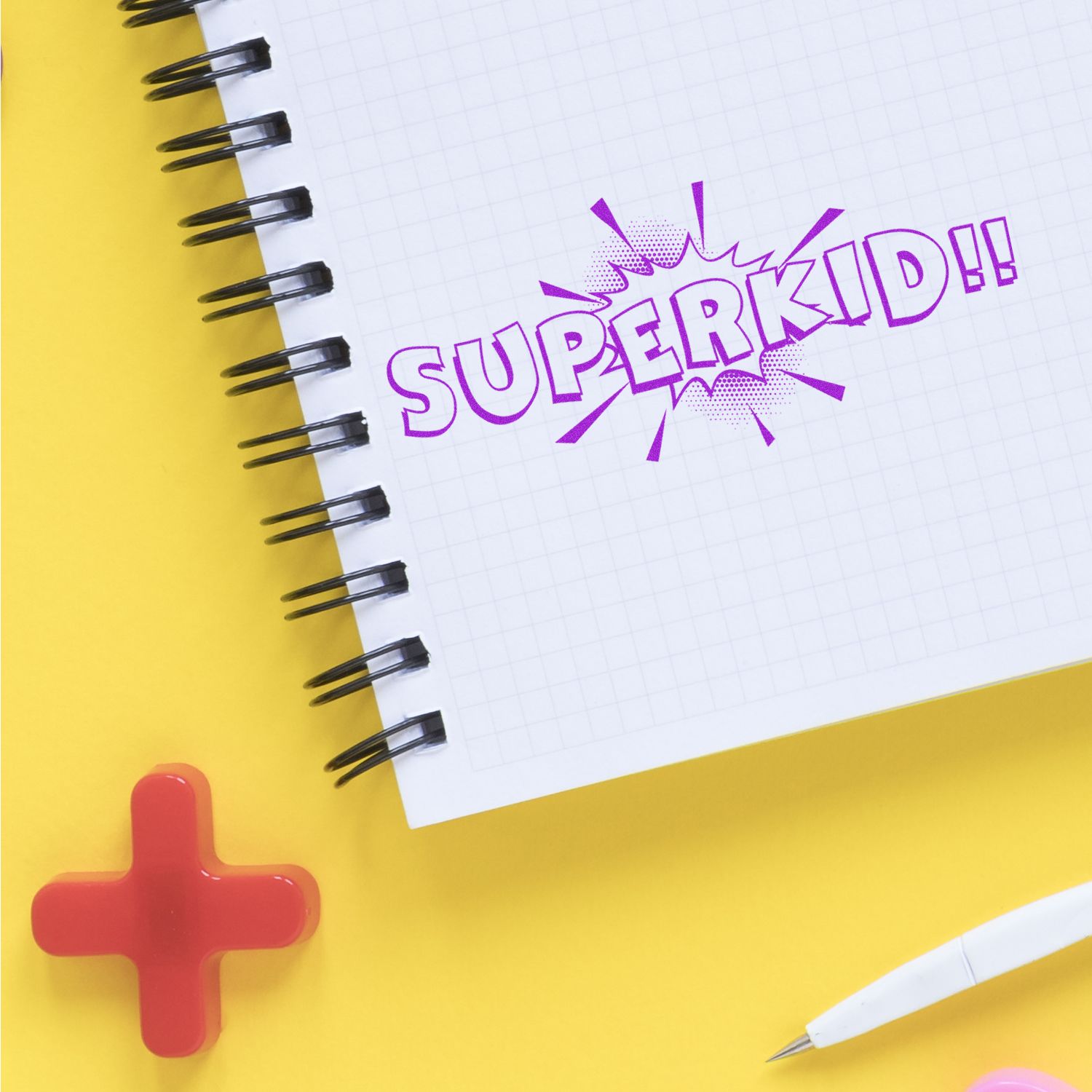 Colorful spiral notebook with purple SUPERKID!! comic text on a yellow background, featuring a pencil and red plus symbol.