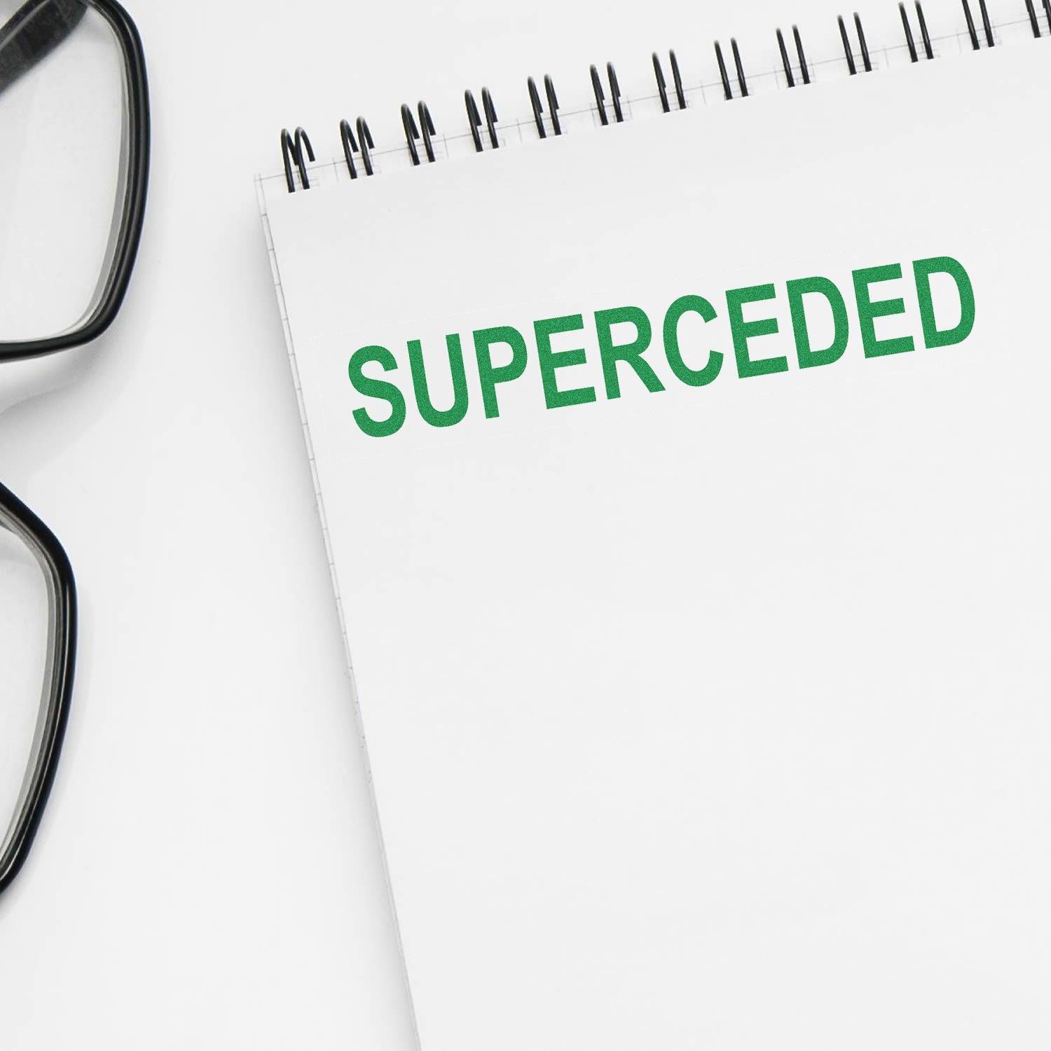 White notebook with green SUPERCEDDED text beside eyeglasses on a clean desk.