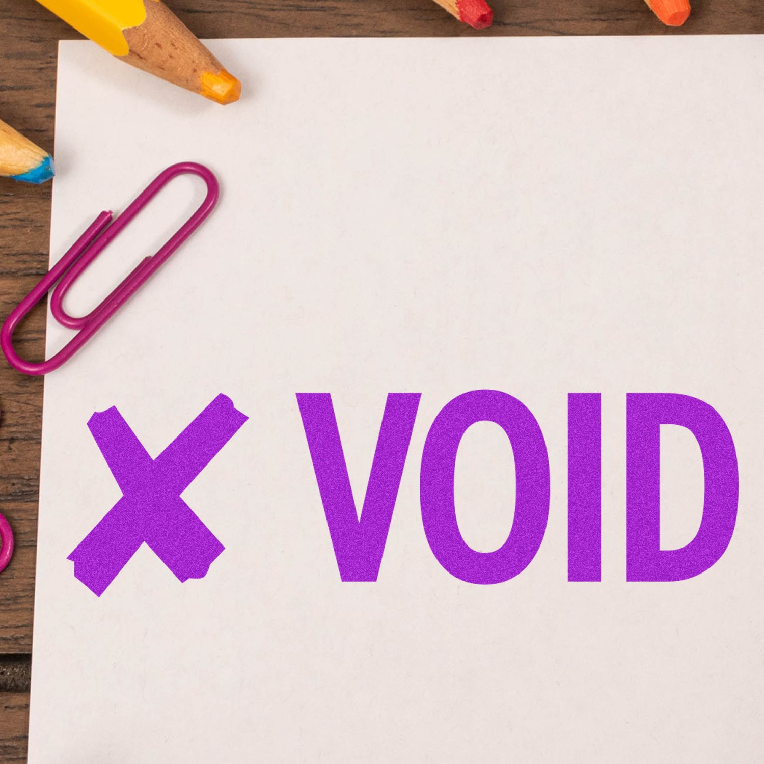 Blank paper marked VOID with a purple paperclip, colored pencils, and office stationery on a wooden desk.