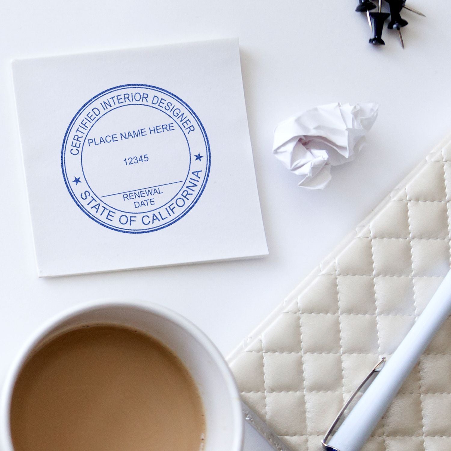 Interior designer workspace flat lay with California certified interior designer stamp, coffee cup, notebook, pen, and crumpled paper on a white desk.