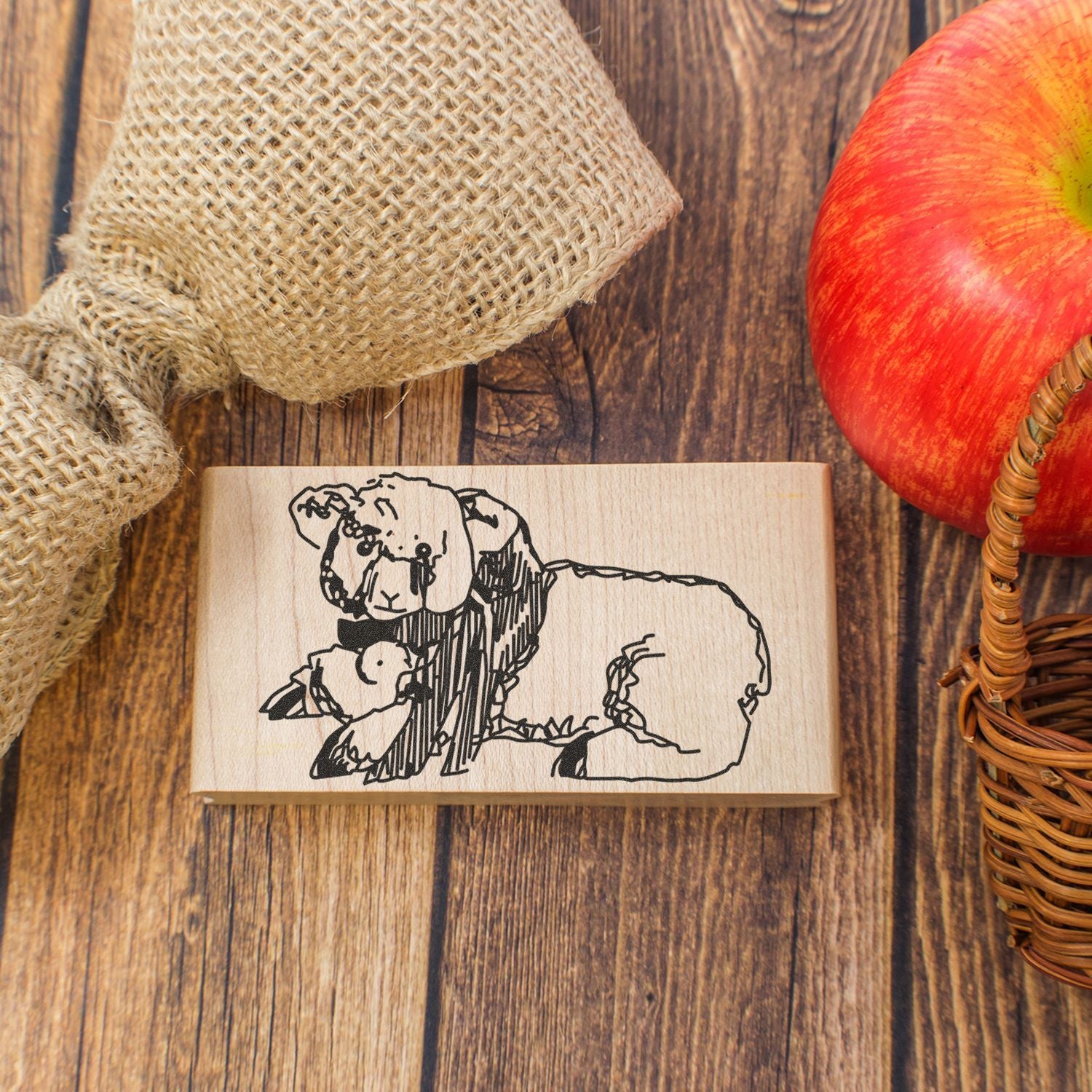 Rustic wooden sheep stamp with lamb illustration on a farmhouse-style wooden table, styled with burlap, an apple, and a woven basket.
