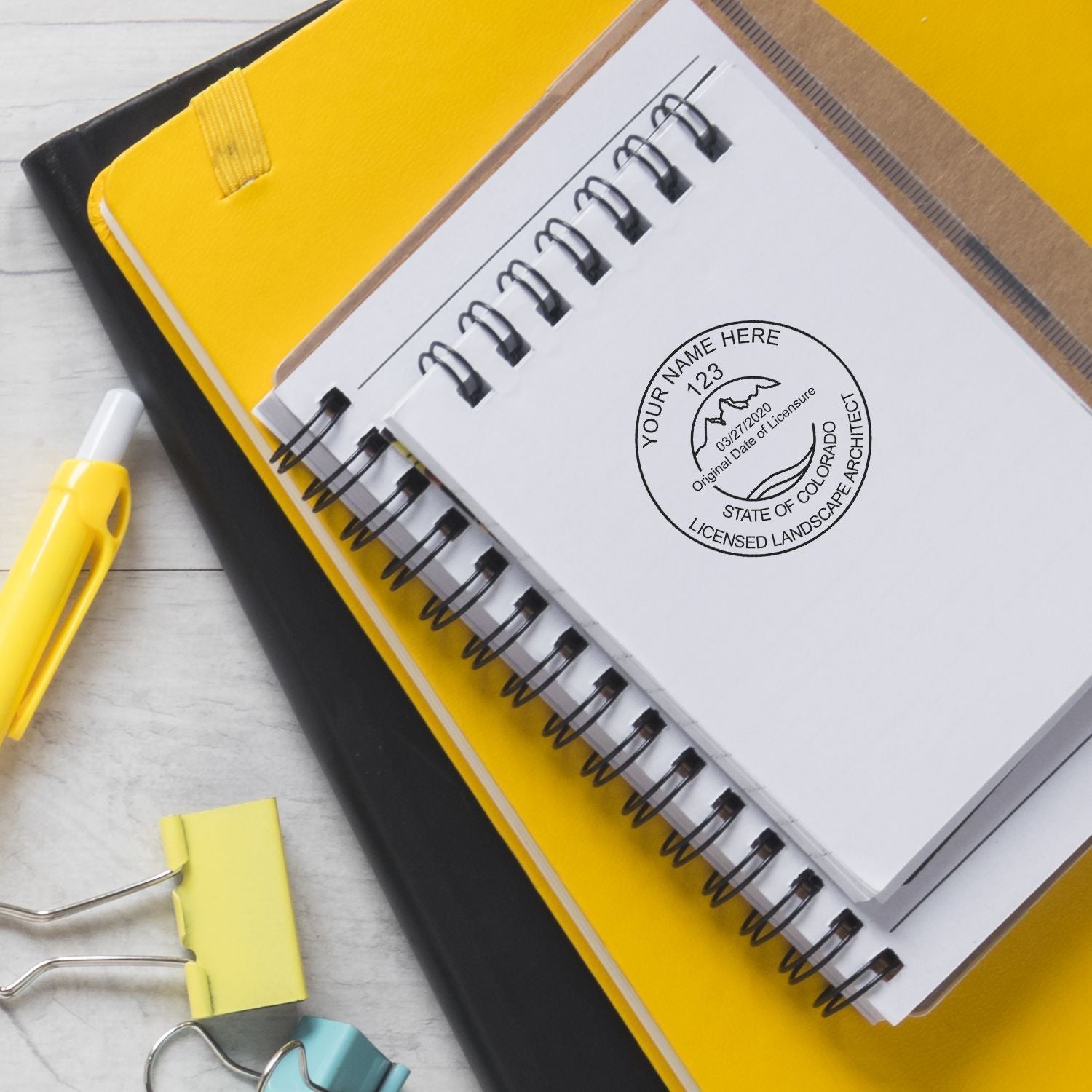 Spiral notebook with Colorado licensed landscape architect stamp on a yellow folder, with binder clips and pen on a desk.