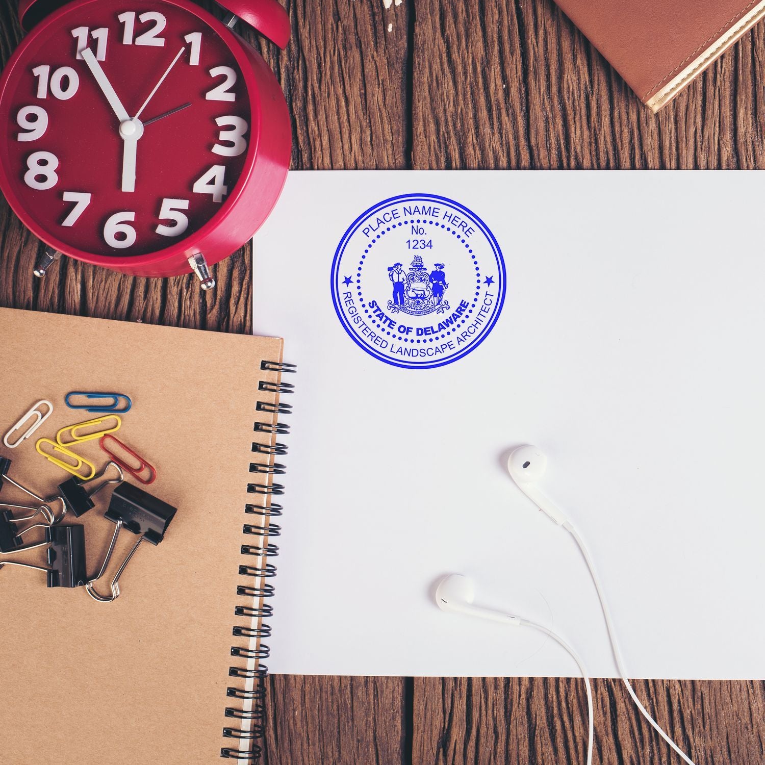 Flat lay of office desk with red alarm clock, notebook, paper clips, earbuds, and Delaware registered landscape architect seal on white paper.