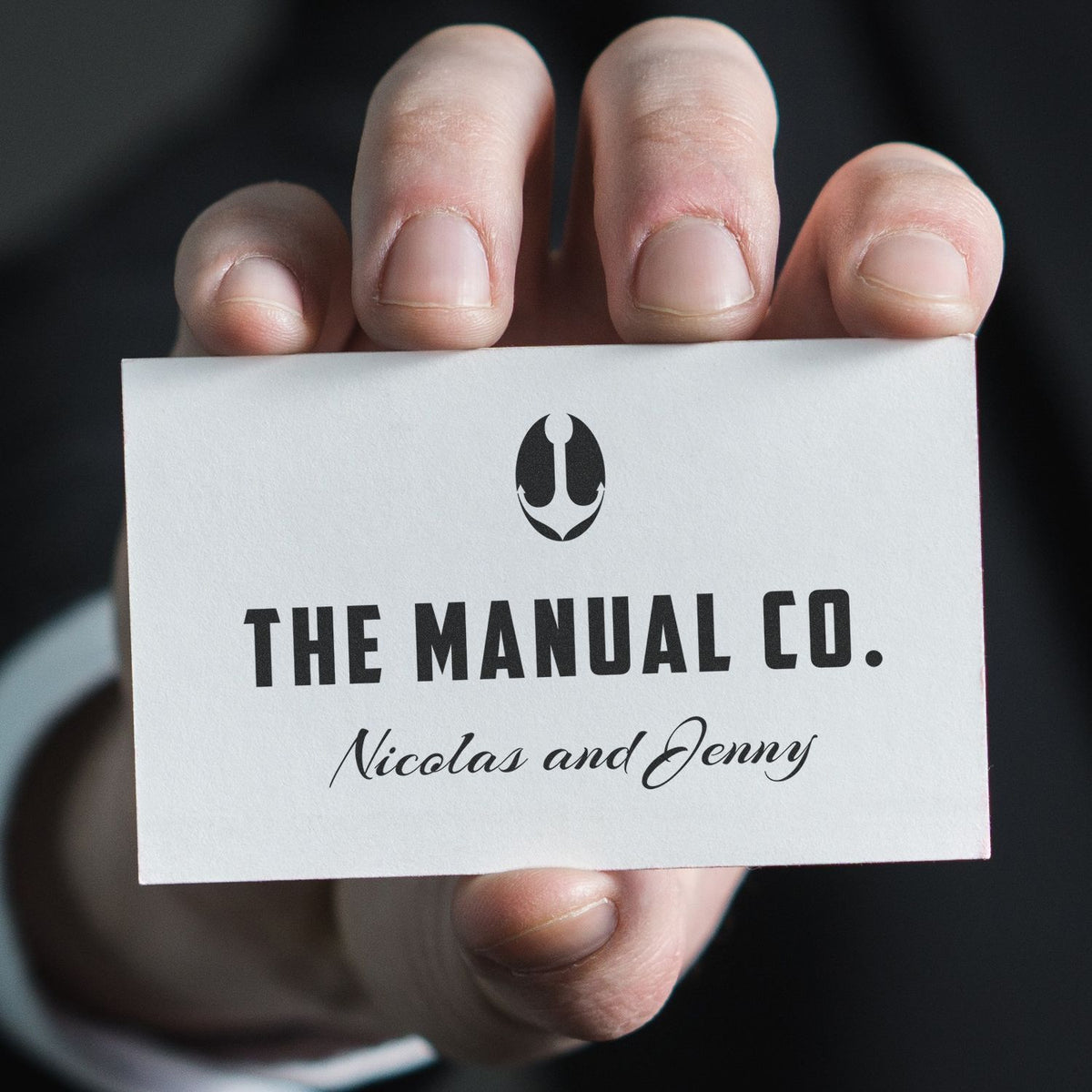 Hand holds a printed card with flame logo and text 'The Manual Co.' plus 'Nicola and Jenny' names, branding cover design for gift.