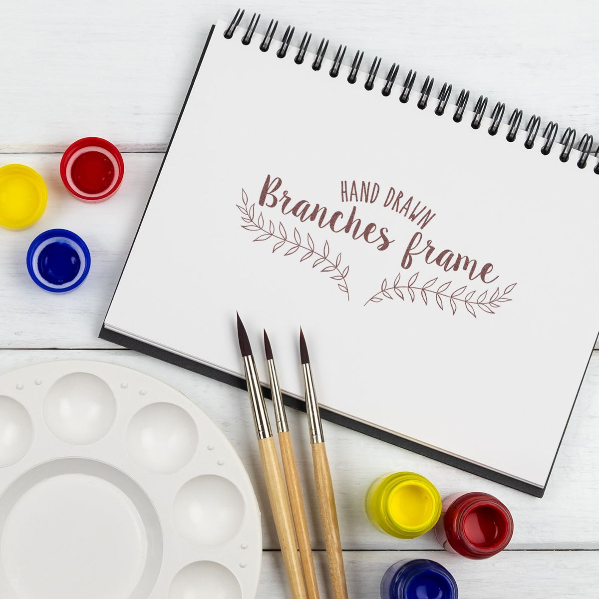 Hand-drawn branches frame sketchbook with paintbrushes, watercolor palette, and paint pots on a white wooden background.