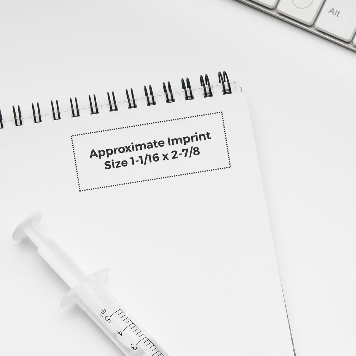 Notebook and syringe on a white desk with Approximate Imprint Size 1-1/16 x 2-7/8 text.