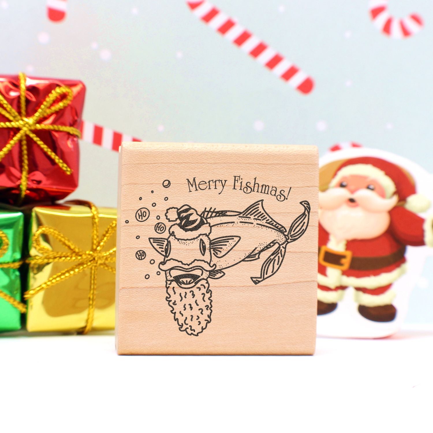Wooden Merry Fishmas holiday stamp with fish Santa design, festive Christmas gifts and Santa decoration in background.