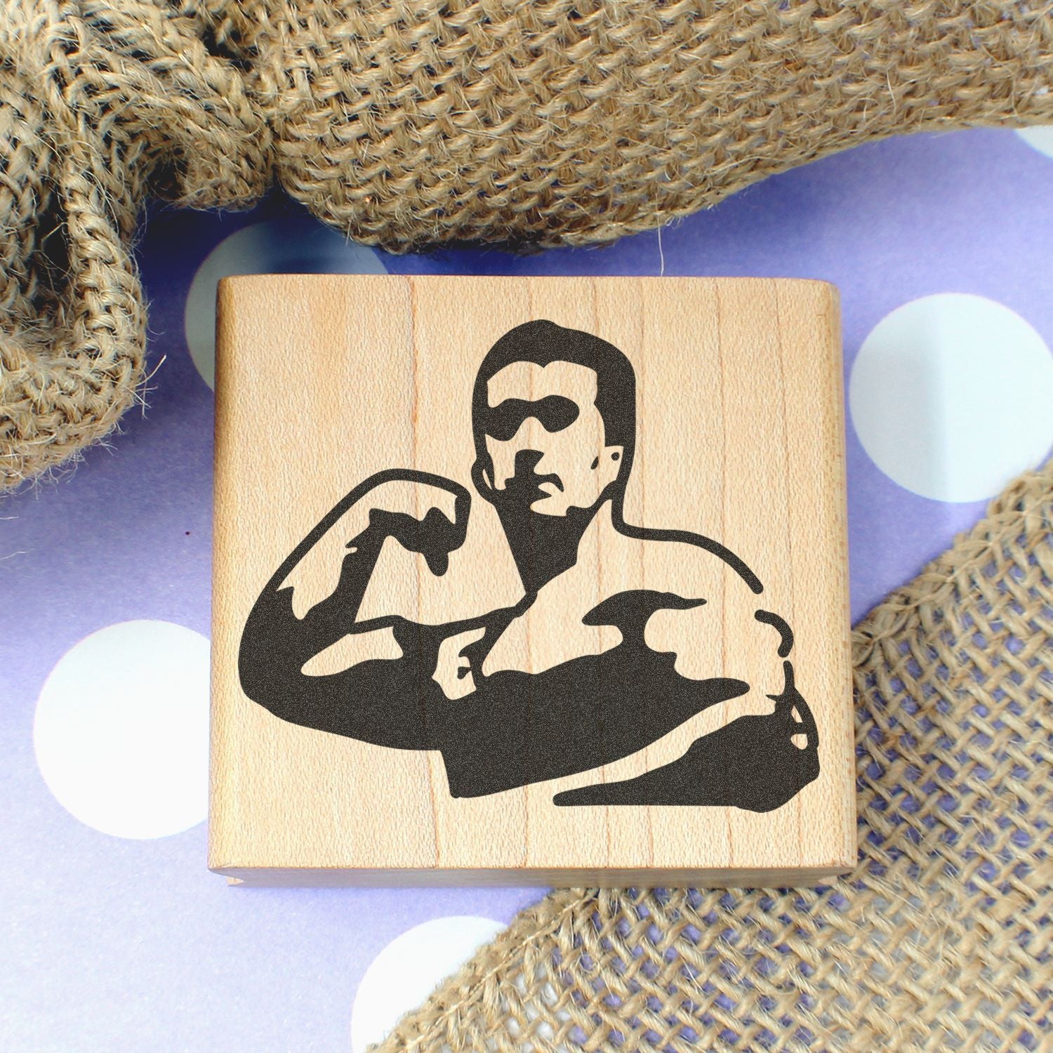 Wooden rubber stamp with flexing bodybuilder illustration on a purple polka dot background with burlap fabric accents