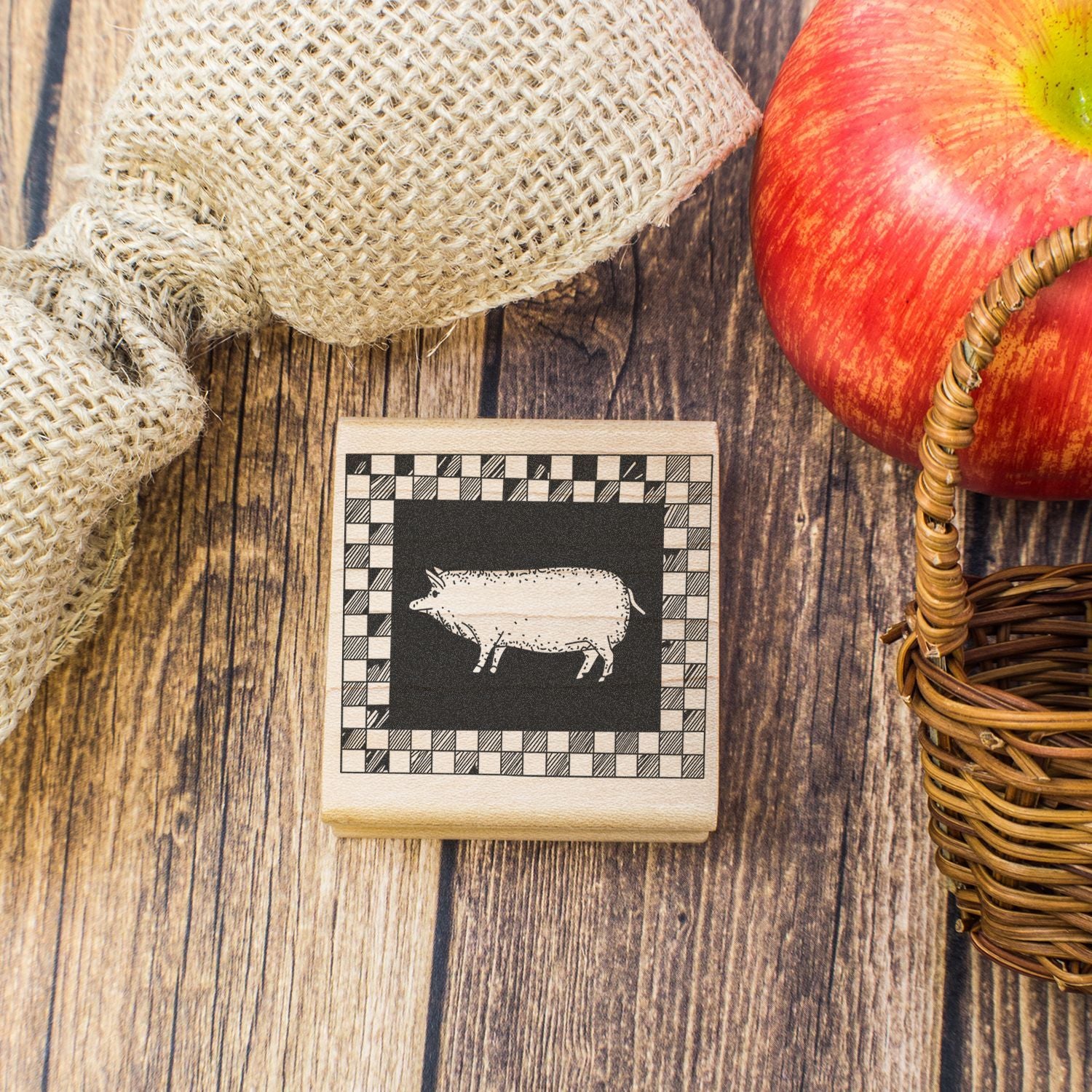 Rustic farmhouse decor with pig print wooden block, burlap sack, red apple, and wicker basket on a wooden background