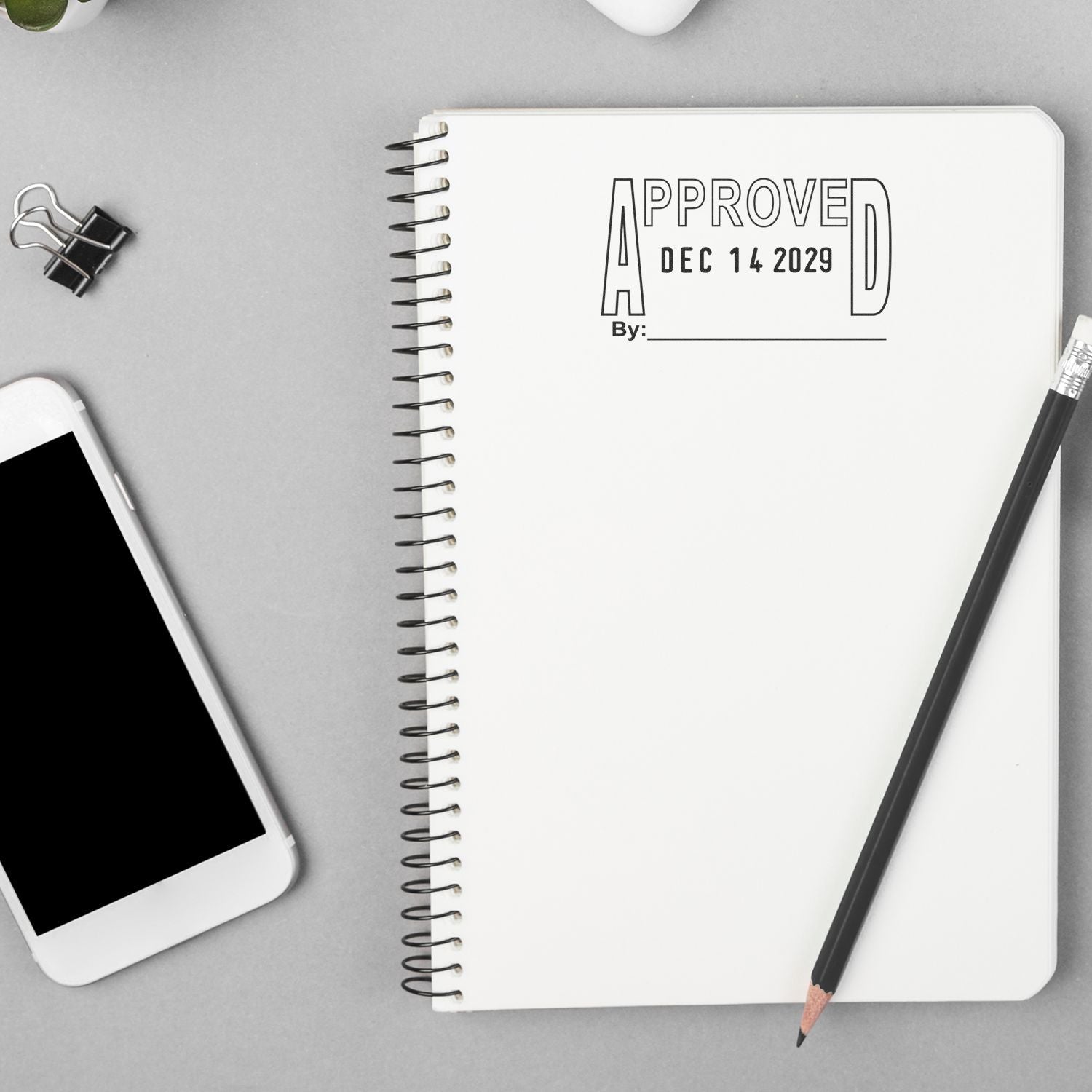 Minimal office desk flat lay with spiral notebook stamped Approved, smartphone, pencil, and binder clip on gray background.
