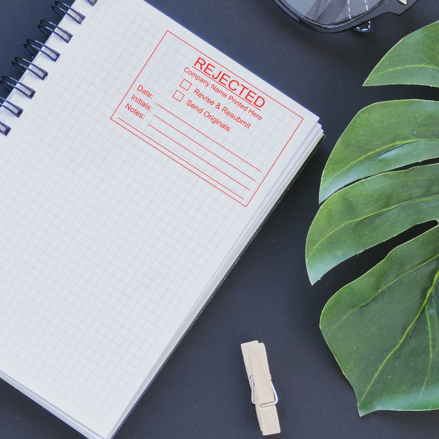 Rejection stamped notebook on black desk with green leaves, eyeglasses, and clothespin, symbolizing business approval failure and document review.