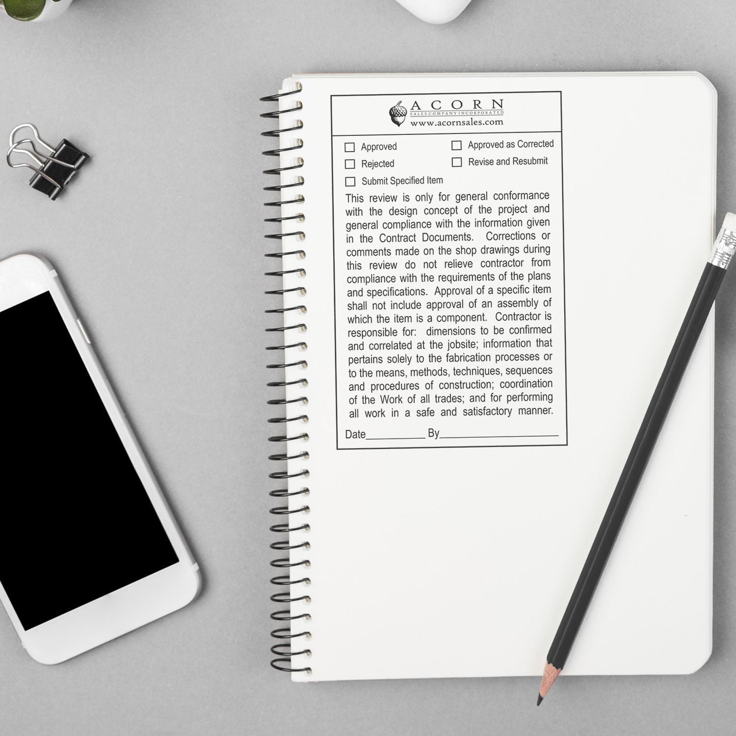 Top view of spiral notebook with project approval form, pencil, smartphone, and binder clip on a gray desk.