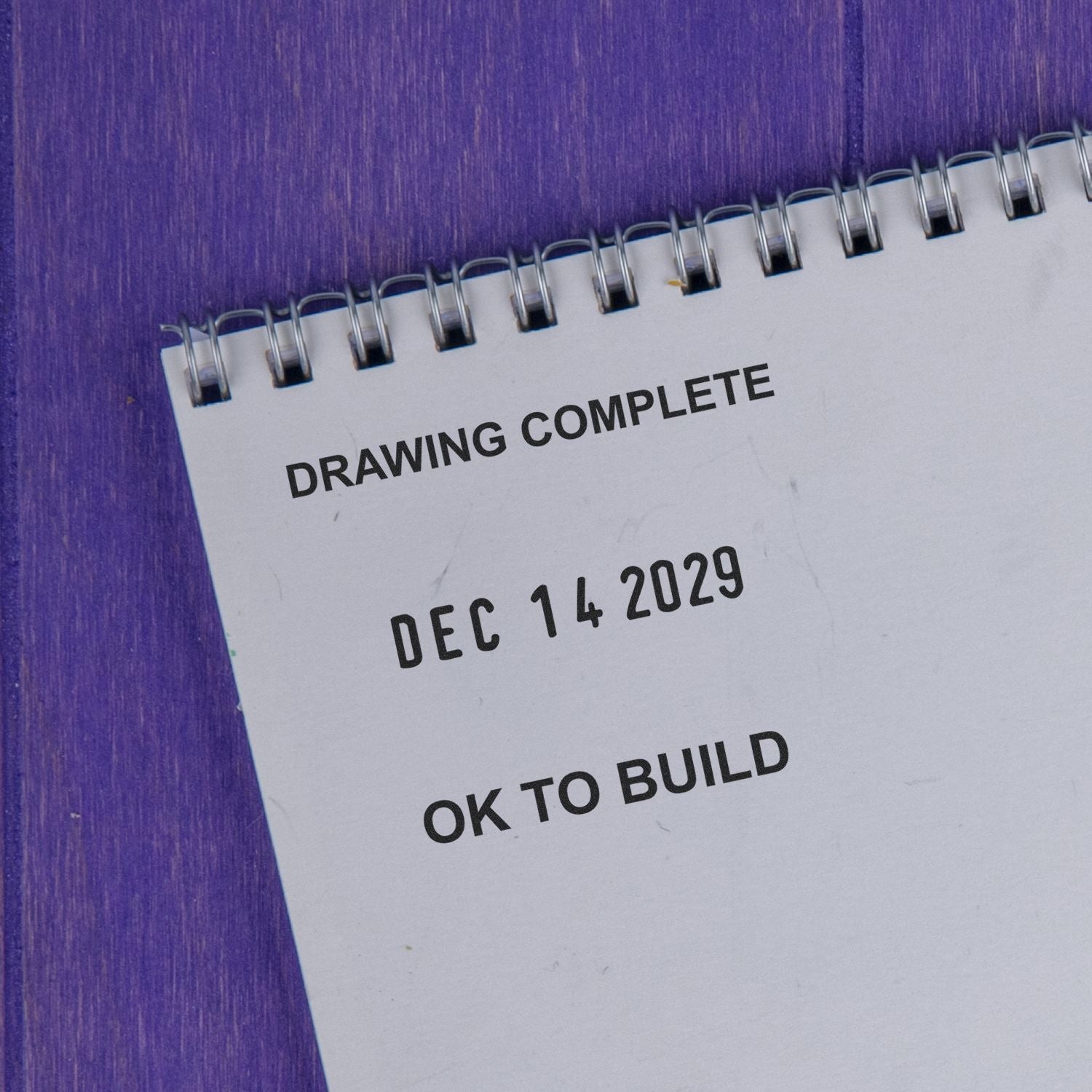 Spiral notebook page reading Drawing Complete, Dec 14 2029, and OK to Build on a purple background.