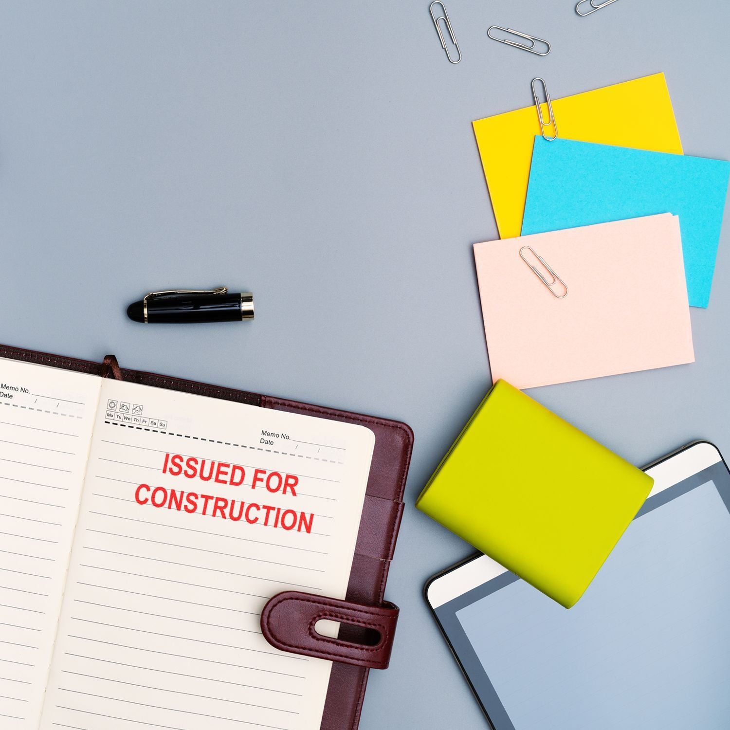 Construction project planning desk with notebook labeled Issued for Construction, colorful sticky notes, pen, paper clips, and smartphone on a gray background.