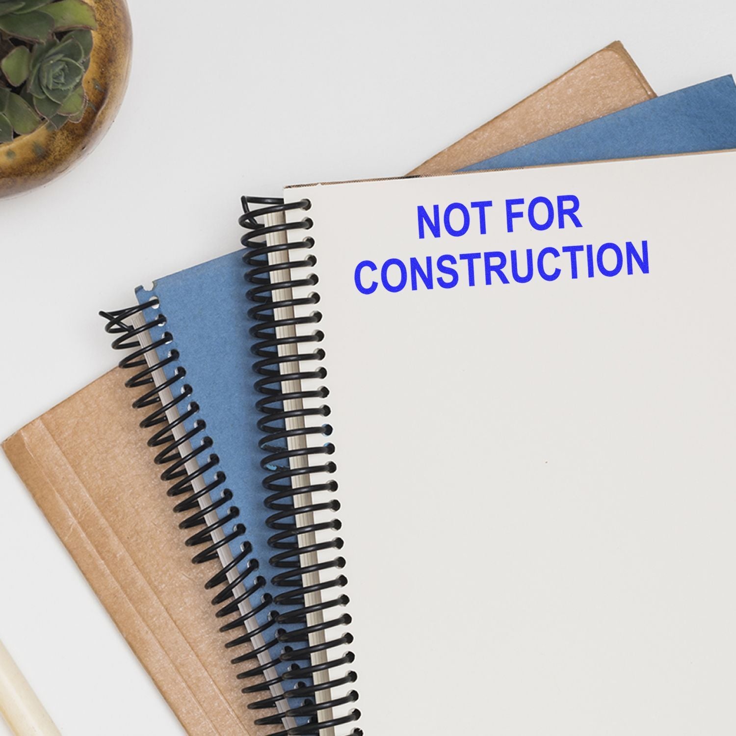 Spiral notebook with Not For Construction text on a desk with stationery and a succulent plant.