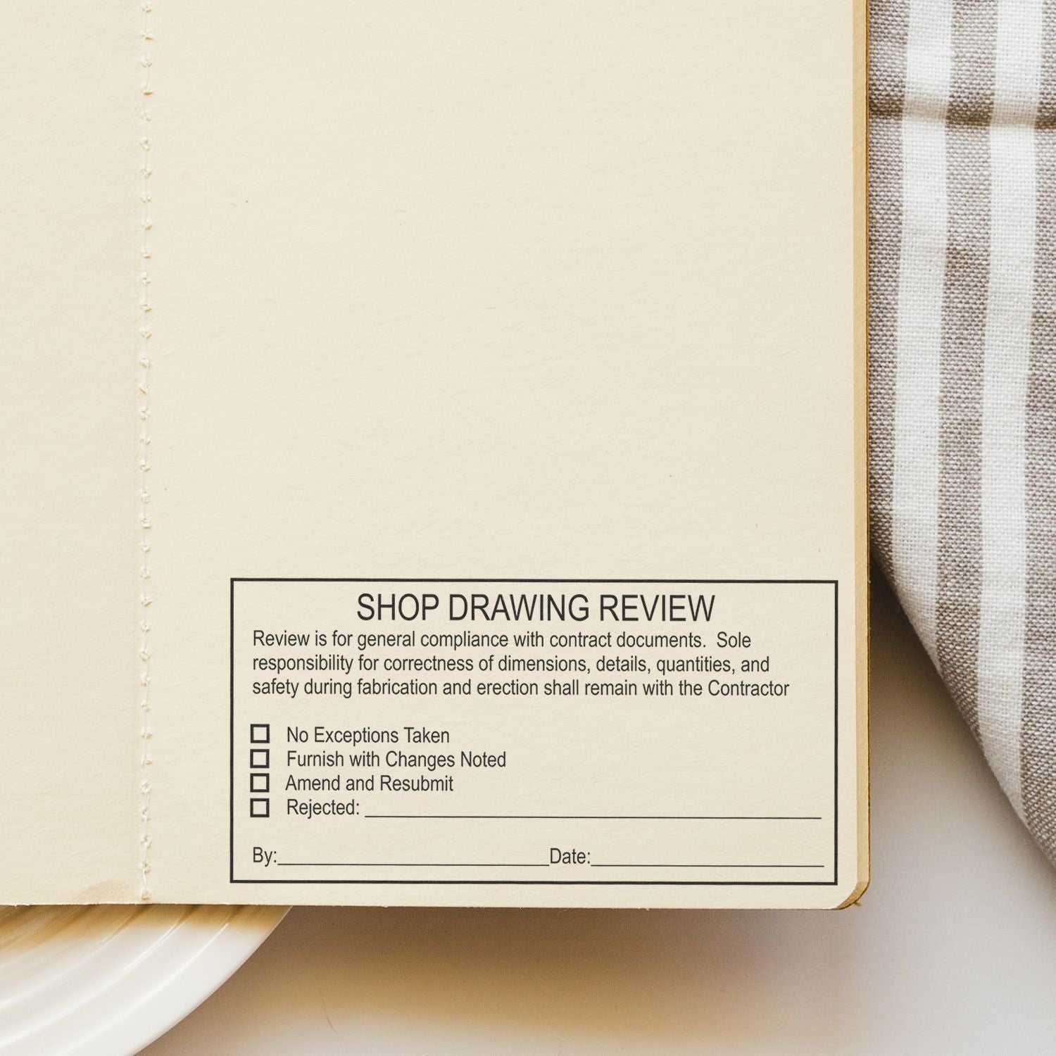 Close-up of an open notebook page with a shop drawing review stamp and plaid fabric background.