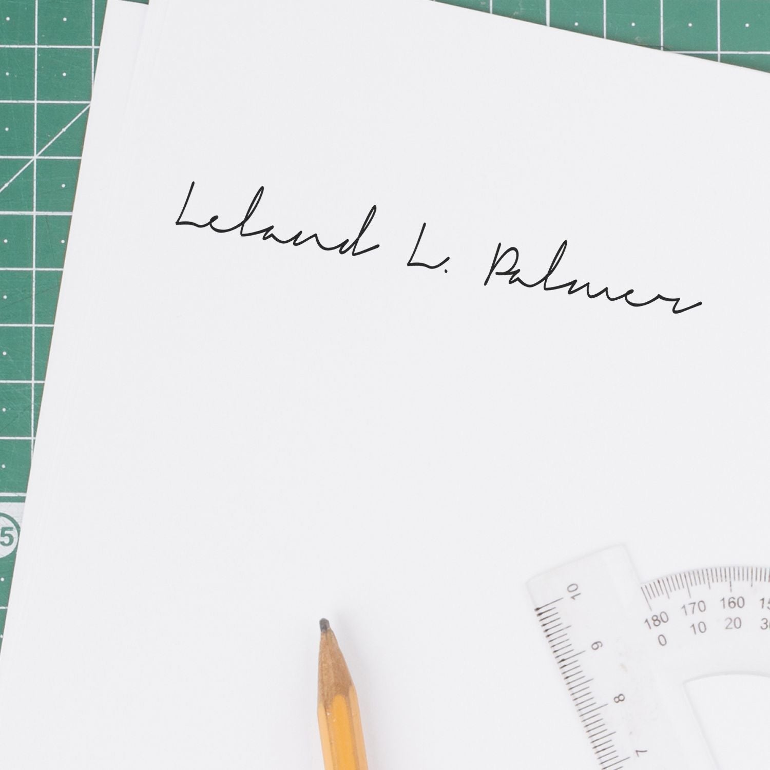 Handwritten signature on white paper with pencil and ruler on a green cutting mat.